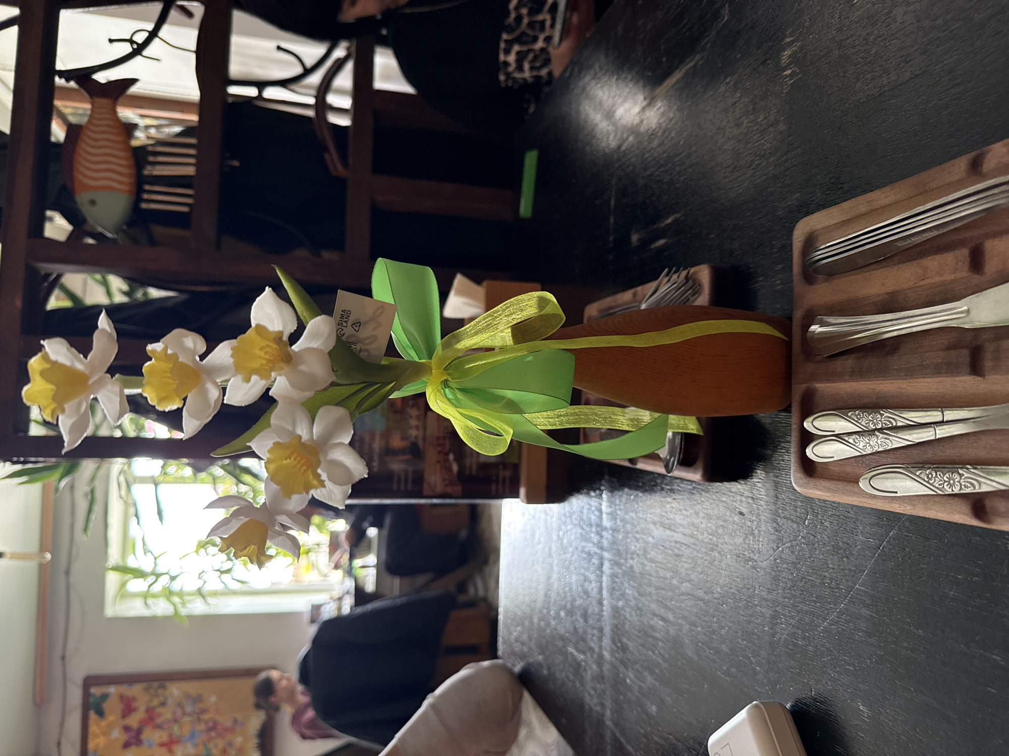 The image shows a table arrangement with a wooden vase containing white and yellow narcissus flowers tied with a green and yellow ribbon. A tag is attached to the vase. To the right of the vase, wooden trays hold silverware. In the background, there is a wooden shelf with a decorative fish. People are visible in the blurred background, with a window and artwork also present. The location is potentially Ust-Koksa, Russia.