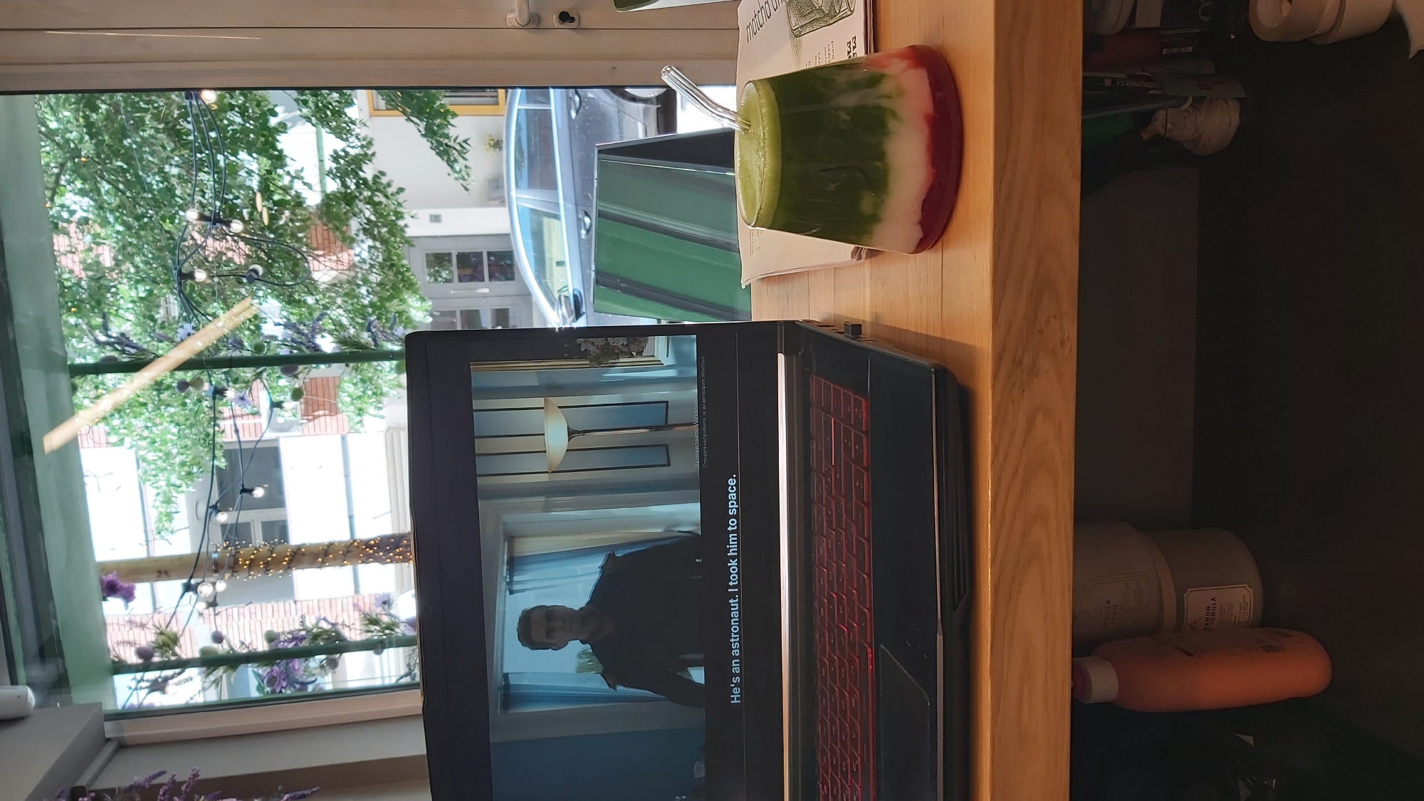 The image shows a laptop displaying a scene with subtitles "He's an astronaut. I took him to space.", next to a glass of layered green and red drink with a straw. The scene is set indoors, with a window view of a street in Sofia, Bulgaria. The street is lined with buildings, trees, and decorative lights, and a car is visible outside. The laptop and drink are on a wooden surface, which seems to be part of a shelving unit filled with various items including bottles, tape rolls, and packaging.