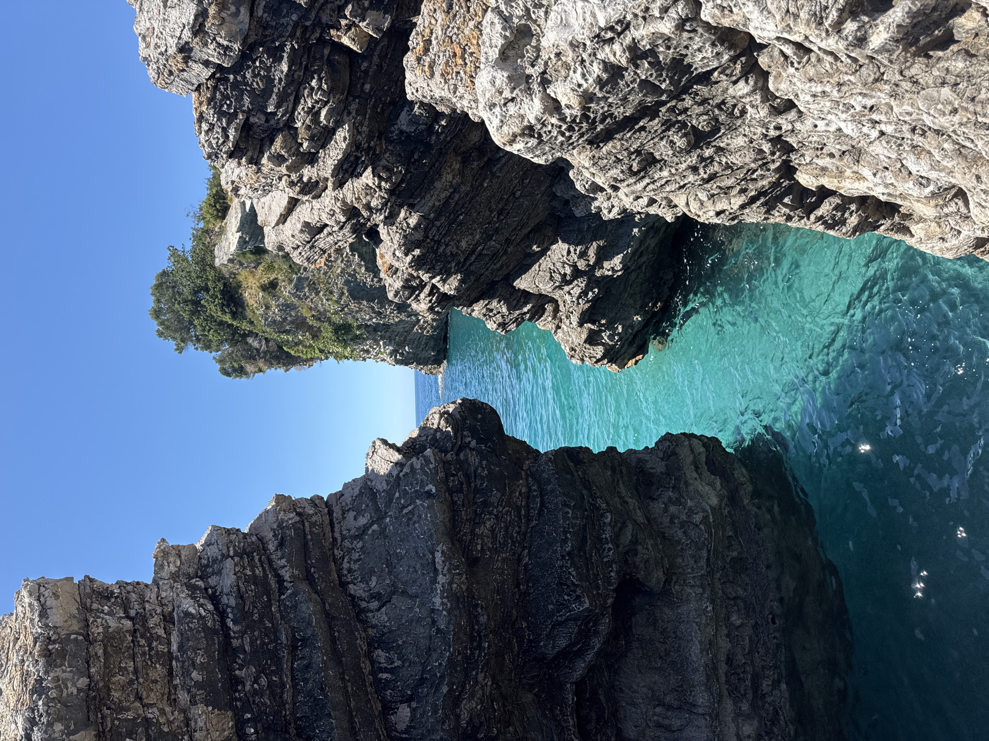 This image presents a stunning natural marine landscape, captured from a perspective looking out through a narrow opening between large, rugged rock formations. On both sides, imposing cliffs dominate the foreground and midground, showcasing layers of grey and brown rock with intricate textures, cracks, and weathered surfaces. Sunlight illuminates the upper sections of these rocky structures, creating strong contrasts and casting shadows that accentuate their rough, ancient appearance.
Between the rock formations, the exceptionally clear sea is a prominent feature, displaying vibrant shades of turquoise and blue. Near the cliffs, the water appears deeper and darker, gradually transitioning to a lighter, almost glowing aqua further out. Subtle ripples and reflections of sunlight dance on the water's surface. In the distance, on the left, a smaller, green-covered landmass with trees indicates a lush coastline. The sky above is a clear, brilliant blue, completely free of clouds.
Near the horizon line, nestled between the distant land and the right-hand rock, a tiny, indistinct white object is visible on the water, possibly a distant boat or swimmer, though it is too small to identify definitively. The scene exudes a sense of tranquility and natural beauty, characteristic of a coastal grotto or a passage through sea cliffs. The bright, sharp lighting indicates a sunny day, likely late morning or early afternoon. The location context provided is Budva, Montenegro, which aligns with the dramatic Adriatic coastal scenery depicted. There is no visible text in the image.