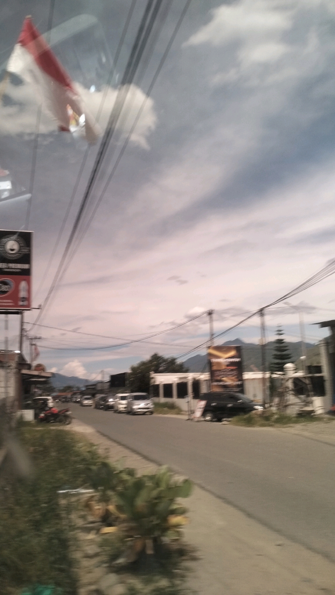 The image depicts a street scene in Nunangantara, Indonesia, under a partly cloudy sky. A red and white flag, likely the Indonesian flag, is visible reflected in a glass surface, possibly a vehicle window. Power lines crisscross the sky, with poles and cables creating a linear pattern. On the left side of the road, a sign with text and graphics is mounted on a pole. Further down the street, several vehicles, including cars and a motorcycle, are present. Buildings line the street, with some appearing to be shops or businesses. In the background, mountains rise against the horizon. Vegetation, including trees and plants, is scattered along the roadside. The overall impression is of a typical urban or semi-urban environment in Indonesia.