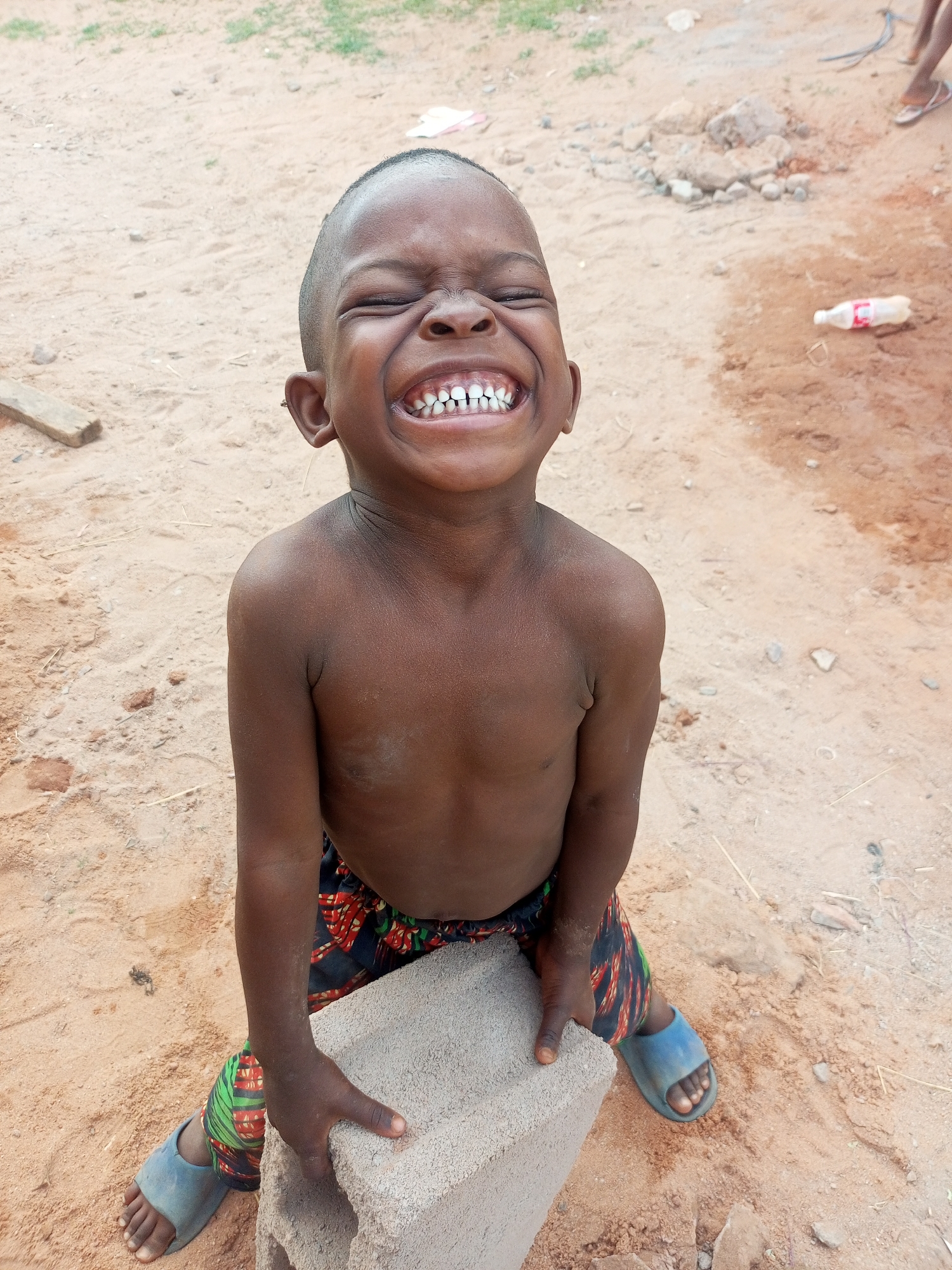 A young, dark-skinned child, appearing to be a boy, is depicted in a close-up outdoor shot, kneeling or squatting on sandy ground. He is shirtless, wearing colorful patterned shorts and light blue slide sandals, and holds a light gray concrete block with both hands.
The child's face is illuminated with a radiant, full-bodied laugh; his eyes are squeezed tightly shut, and his mouth is wide open in a broad grin, revealing a full set of teeth. This expression conveys pure, uninhibited joy.
The setting is an unpaved, sandy or dusty area, likely outdoors during bright daylight. Scattered in the background are small rocks, debris, and a piece of wood on the ground. In the upper right corner, the feet of another person wearing flip-flops are partially visible. An empty, clear plastic bottle with a red cap, resembling a Coca-Cola bottle, lies on the ground further right. The overall impression is one of a simple, natural environment in Tempure, Nigeria, with the child's overwhelming happiness being the focal point. No distinct text is clearly visible, though the implied brand of the bottle is evident.
