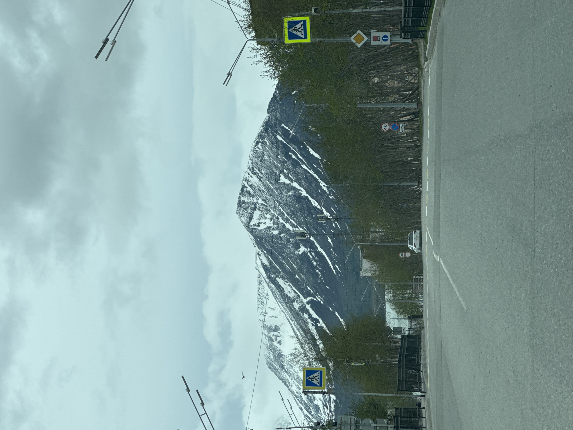 The image depicts a road running alongside a mountain in Apatity, Russia. The mountain is partially covered in snow. A pedestrian crossing sign is visible, along with other traffic signs, including a 40 speed limit sign. Power lines are visible. The sky is overcast. The road is made of asphalt. A few trees with sparse green leaves line the road.