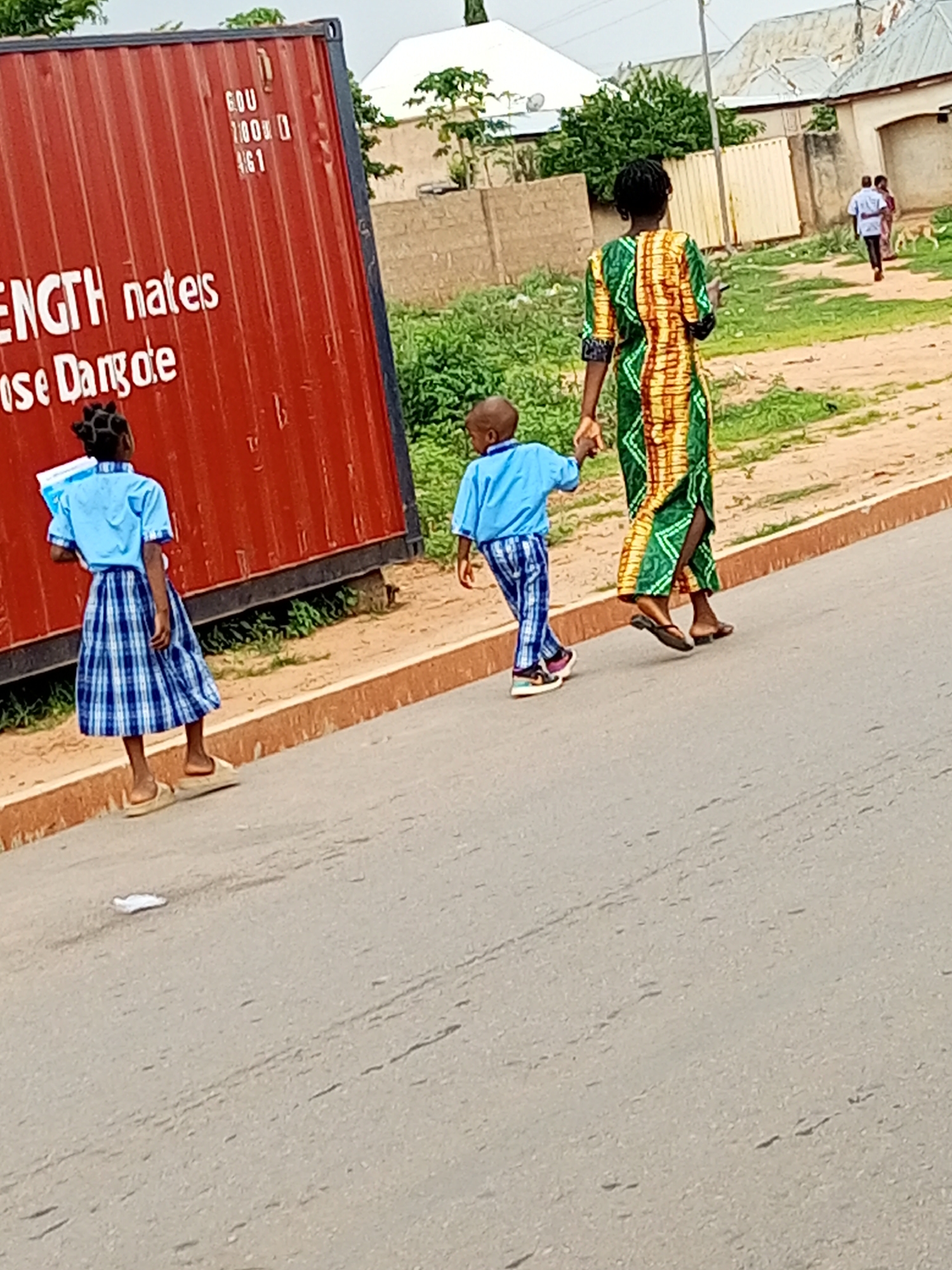 In Tempure, Nigeria, a woman in a vibrantly patterned dress walks hand-in-hand with a young boy wearing a blue shirt and striped pants. Beside them, a young girl in a blue shirt and plaid skirt walks slightly ahead, holding a book. They are walking on a paved road with a brick curb, next to a large red shipping container. In the background, there are buildings with corrugated roofs and scattered greenery. Further in the distance, two other figures and a dog are visible, suggesting a busy street scene.