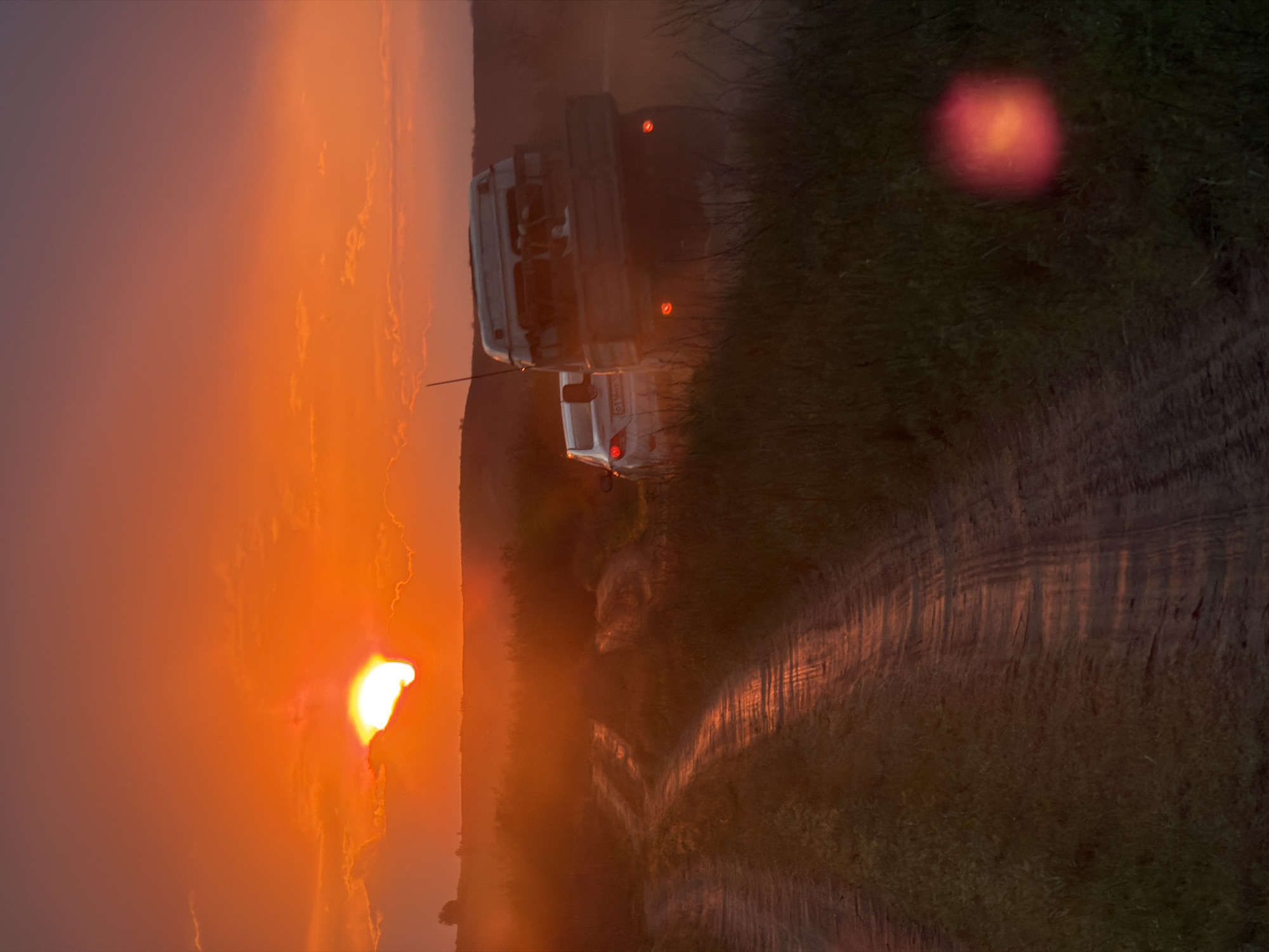 A sideways medium shot depicts a white vehicle, likely a truck or SUV, with its rear lights illuminated, traveling down a dirt path at sunset. The path, flanked by greenery, appears to lead away from the setting sun, which dominates the left side of the frame. The sunset casts a strong orange glow across the sky and landscape, potentially obscuring details of the surrounding environment. The presence of dust suggests the vehicle is traveling on a dry, unpaved surface. The location is Kamyshin, Russia.