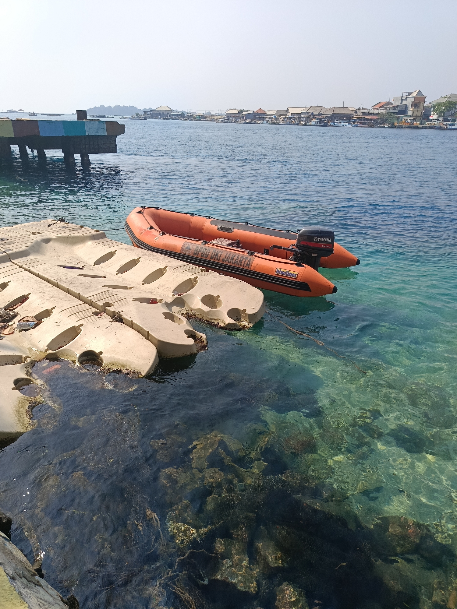 An orange inflatable boat with a Yamaha Enduro outboard motor is tied to a modular floating dock made of beige plastic. The boat has "BPBD DKI JAKARTA" printed on its side, indicating it is associated with the Jakarta Regional Disaster Management Agency. The water is clear and turquoise, revealing rocks and debris below the surface. In the background, a pier with colorful painted sections extends into the water. A hazy skyline of buildings and structures lines the distant shore, suggesting an urban coastal environment within the Special Capital Region of Jakarta, Indonesia. The sky is bright and overcast.