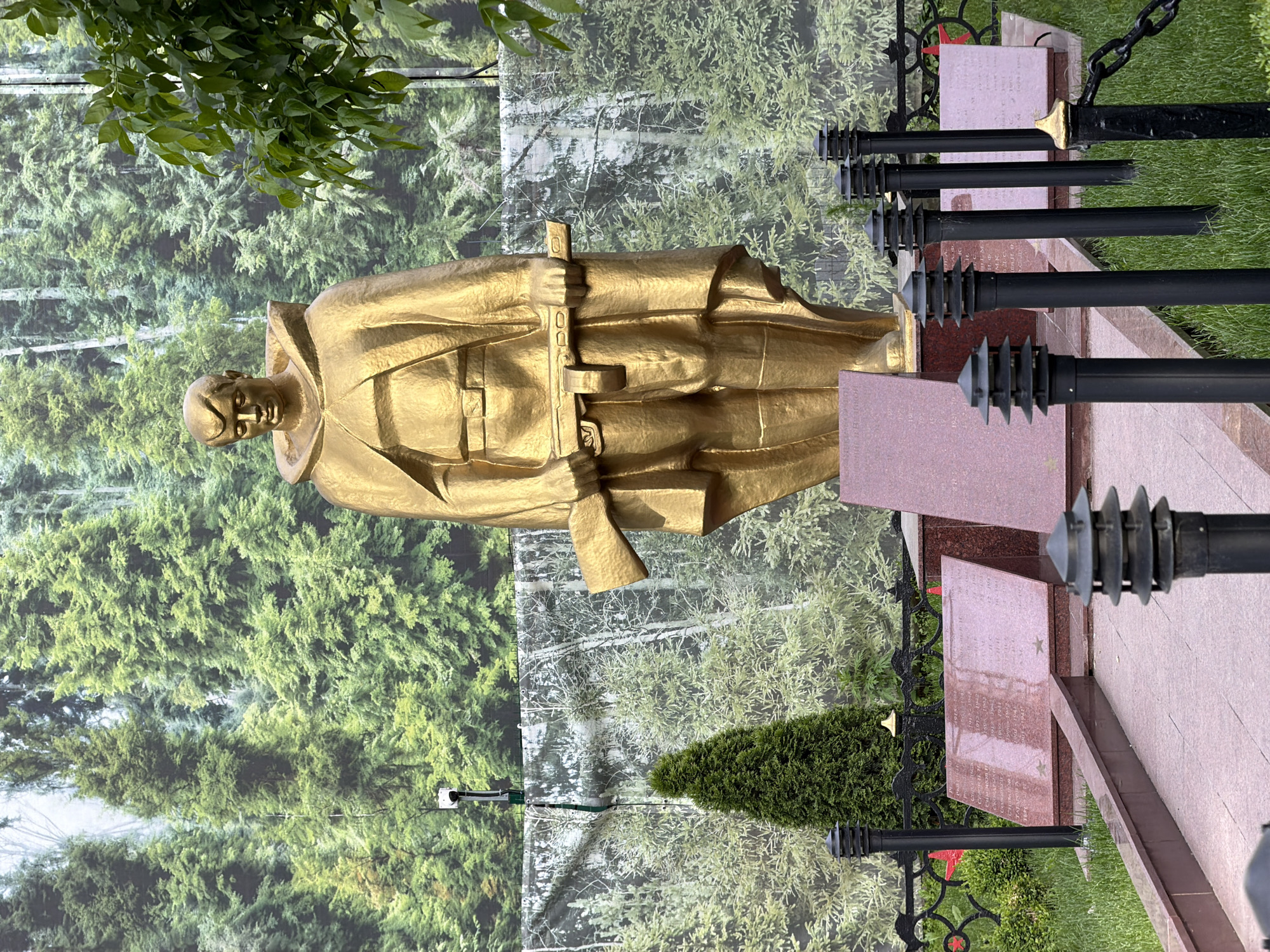 A golden statue of a man in a military uniform stands in front of a backdrop of a forest scene. The statue is centered in the frame and the background is a repeating pattern of green trees. To the right of the statue are black metal posts with cone-shaped tops, separated by lengths of chain. Adjacent to these posts are rose-colored grave markers. The ground is covered in grass, and tree branches are visible in the top left corner of the frame. The location is in the city of Moscow, Russia.