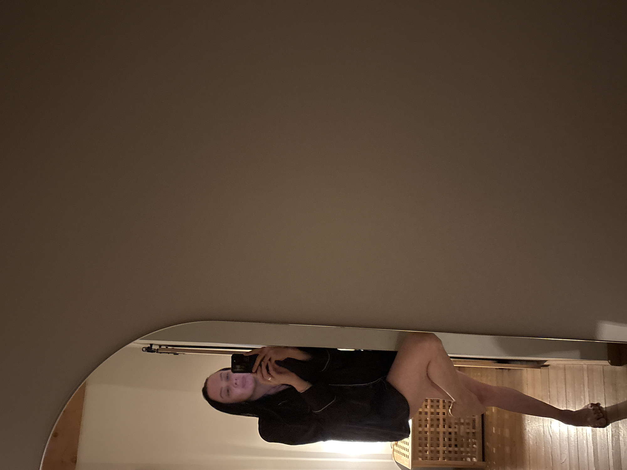 A mirrored image depicts a woman in a dark pajama-like garment, lying down and holding a phone in front of her face. The mirror is oval-shaped with a silver frame. The background visible in the mirror includes a light-colored wall, a wooden slatted chair, and a wooden floor. The top portion of the image is a plain, solid-colored wall. There is no information to confirm the location is Chaikovsky, Russia.