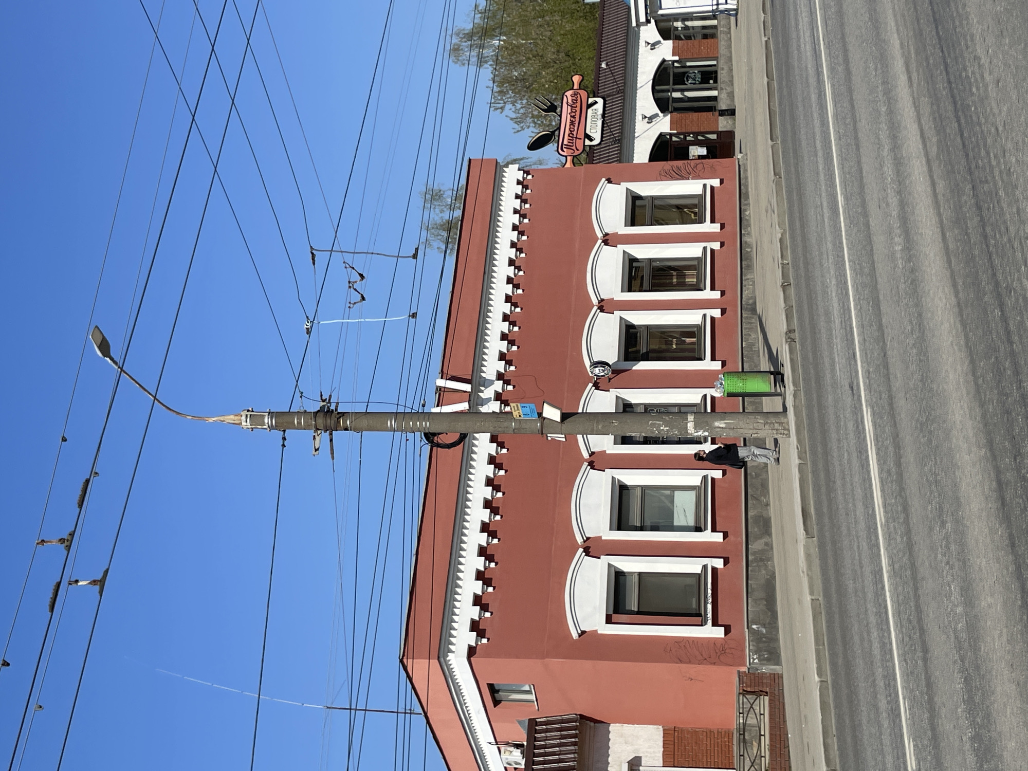 The image captures a building in Izhevsk, Russia, during daylight. The building is primarily red with white architectural trim and several windows. A sign in Russian reads "Пирожковая столовая," indicating a pie shop or canteen. A pedestrian stands on the sidewalk beside the building, near a green trash can. An overhead network of power lines is prominently displayed against a clear blue sky. A street with gray asphalt extends along the side of the building. A light pole stands in front of the building.