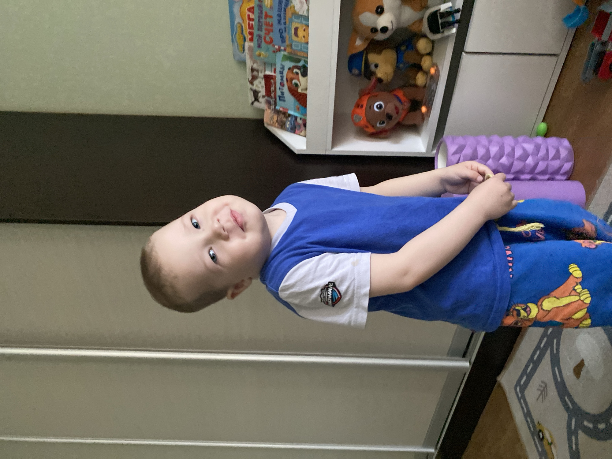 A boy is lying on a white sliding wardrobe in a room. He is wearing a blue T-shirt with white sleeves and blue pants with a cartoon character pattern. A toy shelf is located behind him, containing stuffed animals and children's books, including one with "Paw Patrol" characters. There is a purple foam roller nearby. A section of a rug with road markings is also visible. It is not possible to confirm the location as Sochi, Russia, based on the image alone.