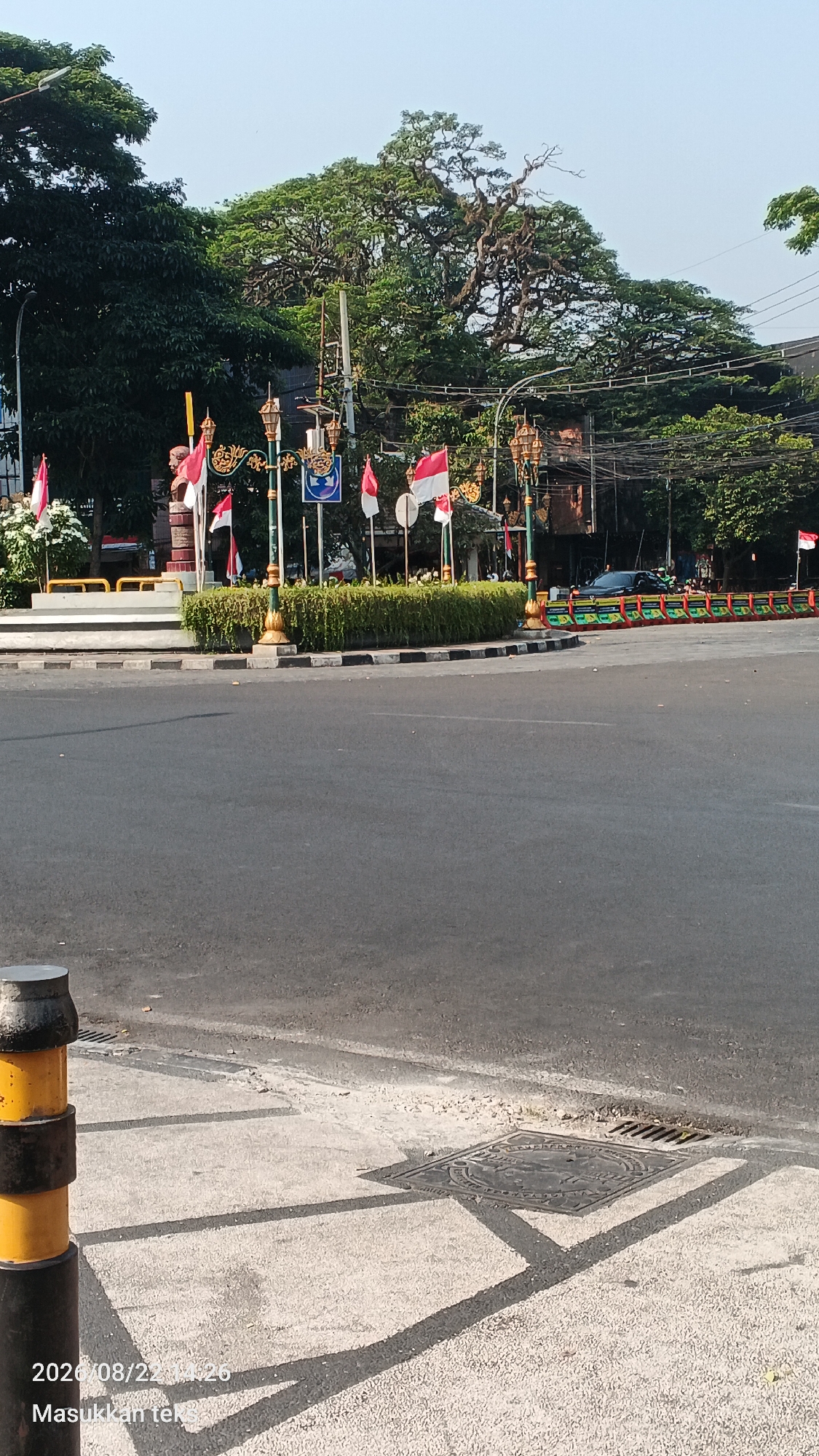 The image shows a street corner in Malang, Indonesia, on August 22, 2020, at 2:26 PM. The scene features a roundabout with several Indonesian flags displayed. There is a bust of a person, likely a historical figure, on a pedestal, surrounded by a hedge and decorative lampposts. The street is paved and appears to be relatively empty of traffic at the moment the photo was taken. In the background, there are trees and buildings. In the foreground, a bollard painted with yellow and black bands is visible, as is a manhole cover. The sky is clear and bright, suggesting a sunny day.