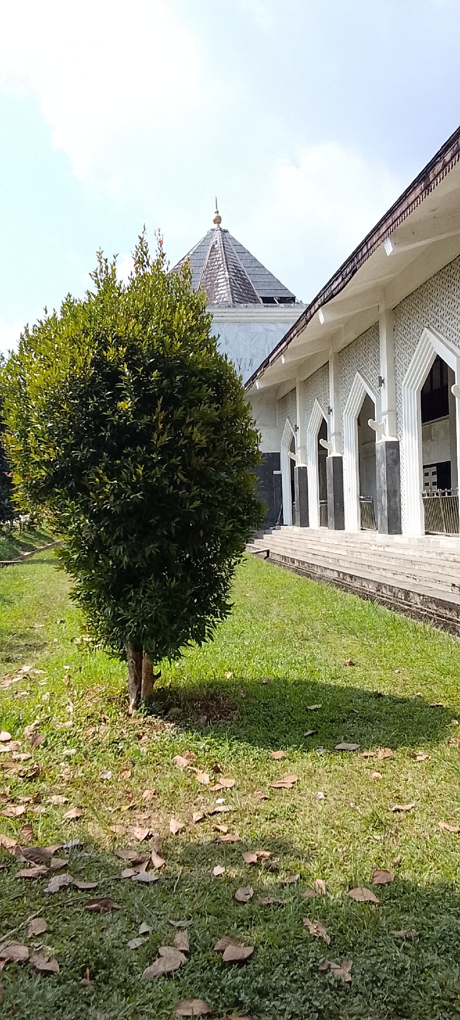 A medium-shot outdoor photograph depicts a lush green tree in the foreground, casting a shadow on the grass. Behind the tree, a mosque with white walls and a gray tiled roof is visible, with its minaret peeking above the building's structure. The mosque features a series of arched doorways and a long, paved walkway leading up to it. The sky is blue with a few scattered clouds. The scene is located in Padang Dalom, Indonesia.