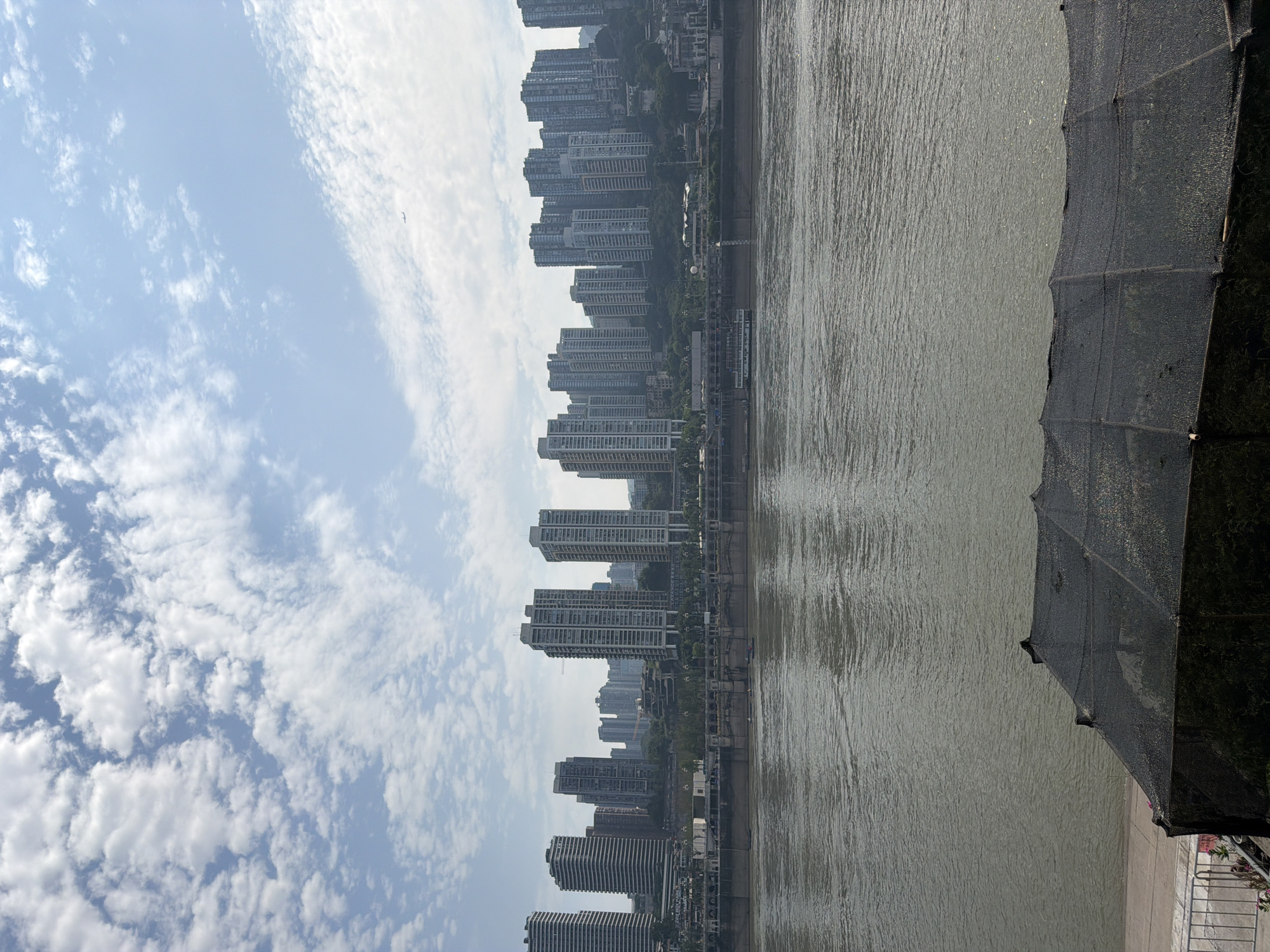 The image shows a wide, murky river in the foreground, with a row of tall, modern buildings lining the opposite bank. The buildings are densely packed, creating a cityscape that stretches across the horizon. A promenade or walkway is visible along the river's edge in front of the buildings, with trees and some architectural elements. In the sky, there are scattered white clouds against a light blue backdrop. A small airplane is visible in the distance, high in the sky. In the lower right corner of the foreground, there is a dark, netted structure, possibly a temporary shelter or enclosure. The scene appears to be a view of the city from across the river, likely in Nan'an District, China, given the architectural style and the presence of a major waterway. The lighting suggests it is daytime, possibly morning or late afternoon, with the sun casting a diffuse light. There are no people clearly visible in the image.