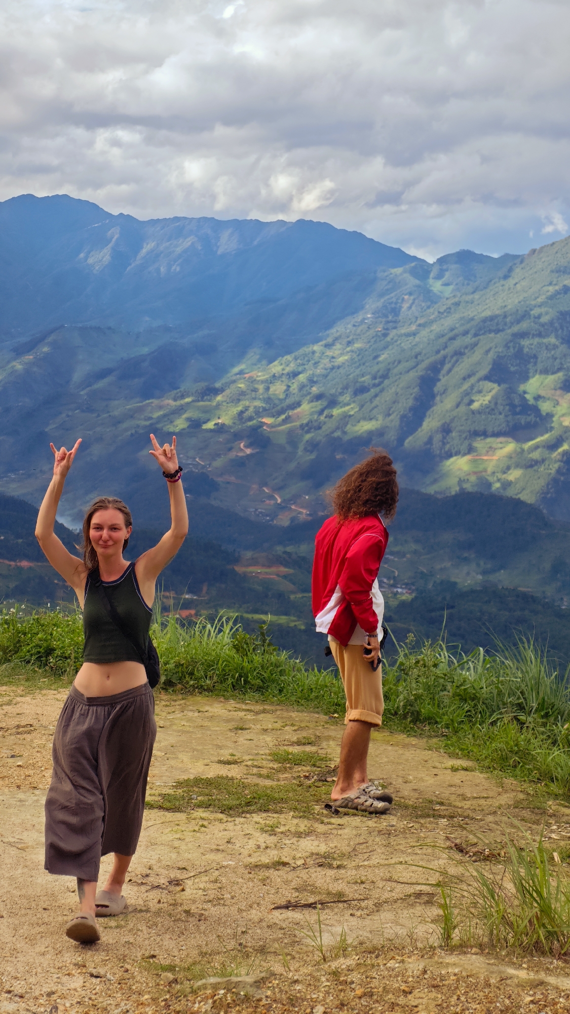 The scene is located in the city of Hanh Phuc, Vietnam, specifically at a mountainous overlook. The sky is predominantly overcast with white and light grey clouds. In the background, multiple layers of lush green mountains extend into the distance, exhibiting varying shades of blue-green haze and featuring visible terraced sections or cultivated areas with winding dirt paths.
In the immediate foreground, two individuals stand on a light brown dirt path interspersed with patches of green grass. On the left, a young adult female faces the viewer, displaying a subtle smile. She wears a dark green sleeveless crop top, dark loose-fitting pants, and light-colored slip-on sandals. Her arms are raised above her head, with both hands forming the "rock on" gesture (index and pinky fingers extended). A dark strap crosses her torso, and multiple dark bracelets adorn her left wrist. A tattoo is visible on her lower left leg.
On the right, a second individual, appearing male, stands with his back mostly to the camera, observing the panoramic mountain vista. He has long, curly reddish-brown hair and is dressed in a red jacket with a white stripe down the sleeves, light beige shorts, and open-toed sandals. A white garment is visible beneath his jacket. He holds a dark-colored smartphone in his left hand.