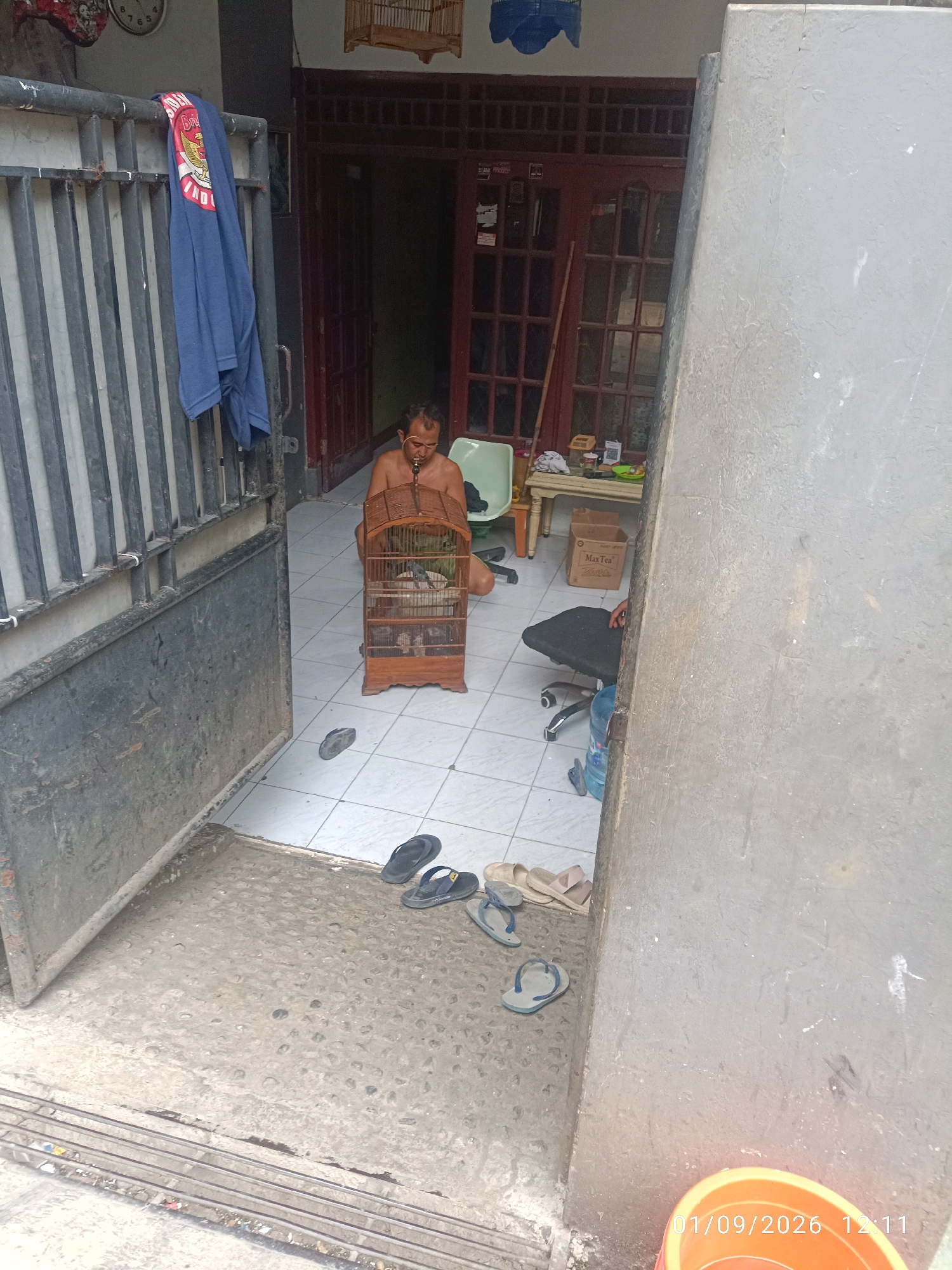 A partially undressed man is sitting on the floor in an open doorway. He is holding a wooden bird cage which contains a bird. In front of him are a pair of black flip-flops. To the left is a metal gate and a dark blue shirt is hanging on the gate. Behind the man, to the right, is a table with items on it and a cardboard box. Further back, there are double doors. To the right of the doorway, a grey wall partially obscures the scene. Scattered on the floor near the doorway are several pairs of flip-flops. A water cooler jug is visible next to a black office chair. A portion of an orange bucket is visible in the lower right corner of the image. A timestamp "01/09/2026 12:11" is visible on the lower right corner.