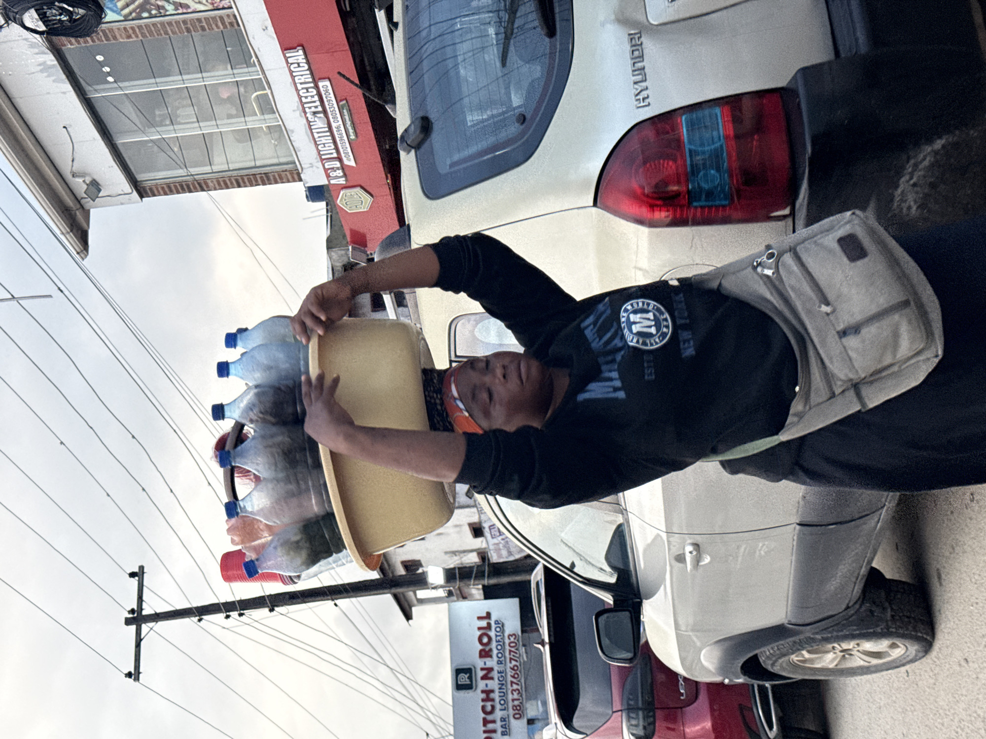 In Ado, Nigeria, a woman carries a yellow basin filled with bottles of beverages on her head. She is standing on a street, with cars parked behind her. A red and white sign for "PITCH-N-ROLL BAR, LOUNGE ROOFTOP" with a phone number is visible in the background. Another building with "A&D LIGHTING ELECTRICAL" signage is also present. The scene appears to be set during daylight with overcast skies.