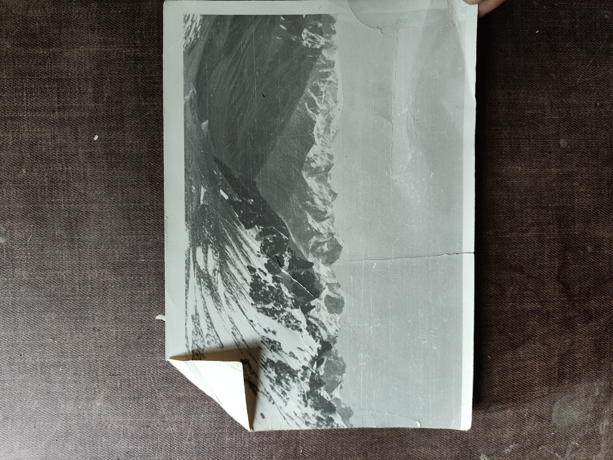 The image shows a vintage black and white photograph of a mountainous landscape. The photograph is slightly creased and torn, with a corner peeled back. The background is a dark, textured surface, possibly fabric or wood. There is no discernible location in the city of Izhevsk, Russia, as the image depicts a natural, mountainous scene.