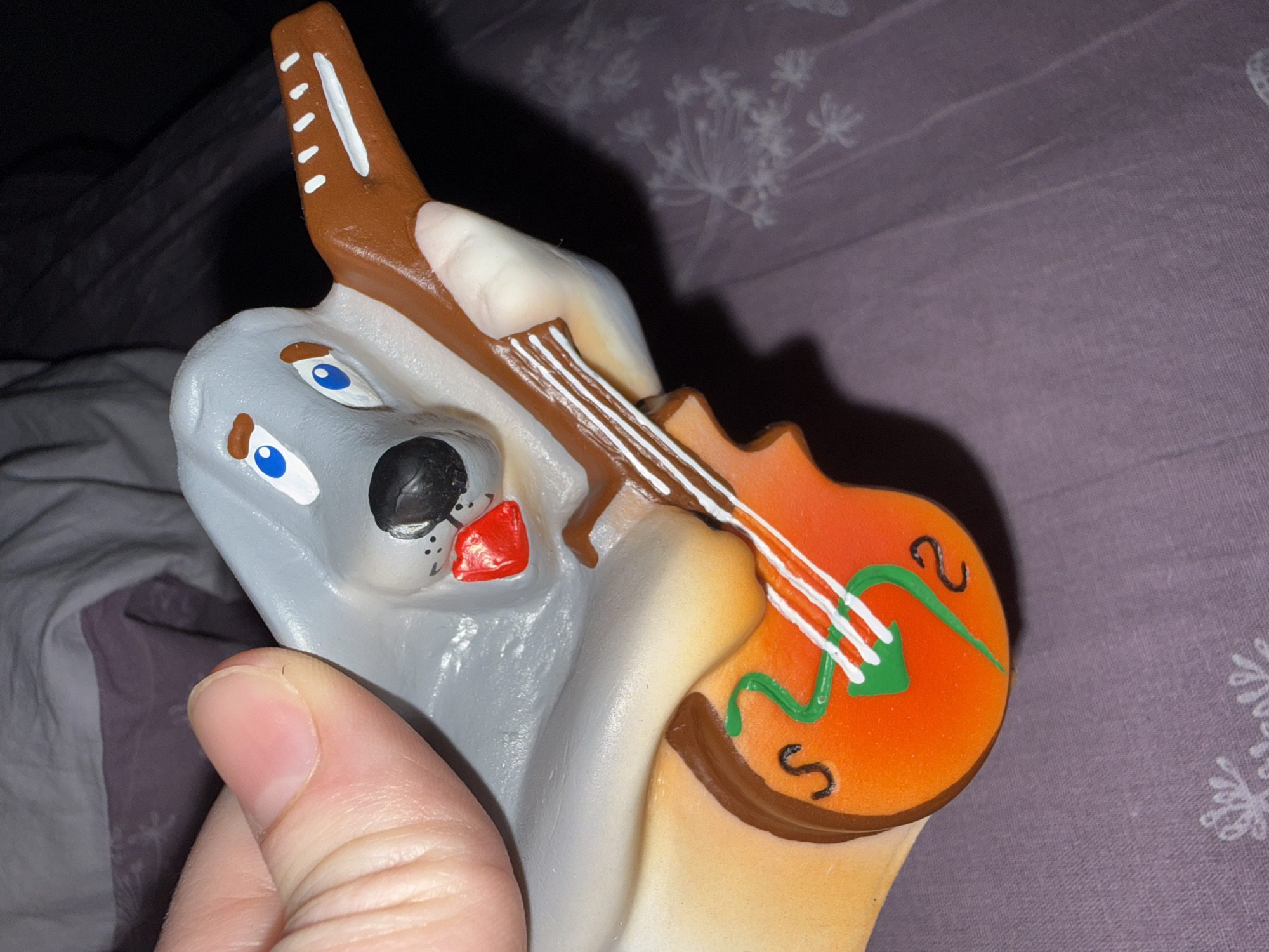 A hand holds a painted ceramic figure of a wolf playing a guitar indoors. The wolf is painted with a grey coat, white underbelly, black nose, red tongue, and blue eyes. The guitar is painted orange with brown trim, white strings, and a green design. The background includes a bed with grey and purple patterned linens. The location and city are not determinable from the image.