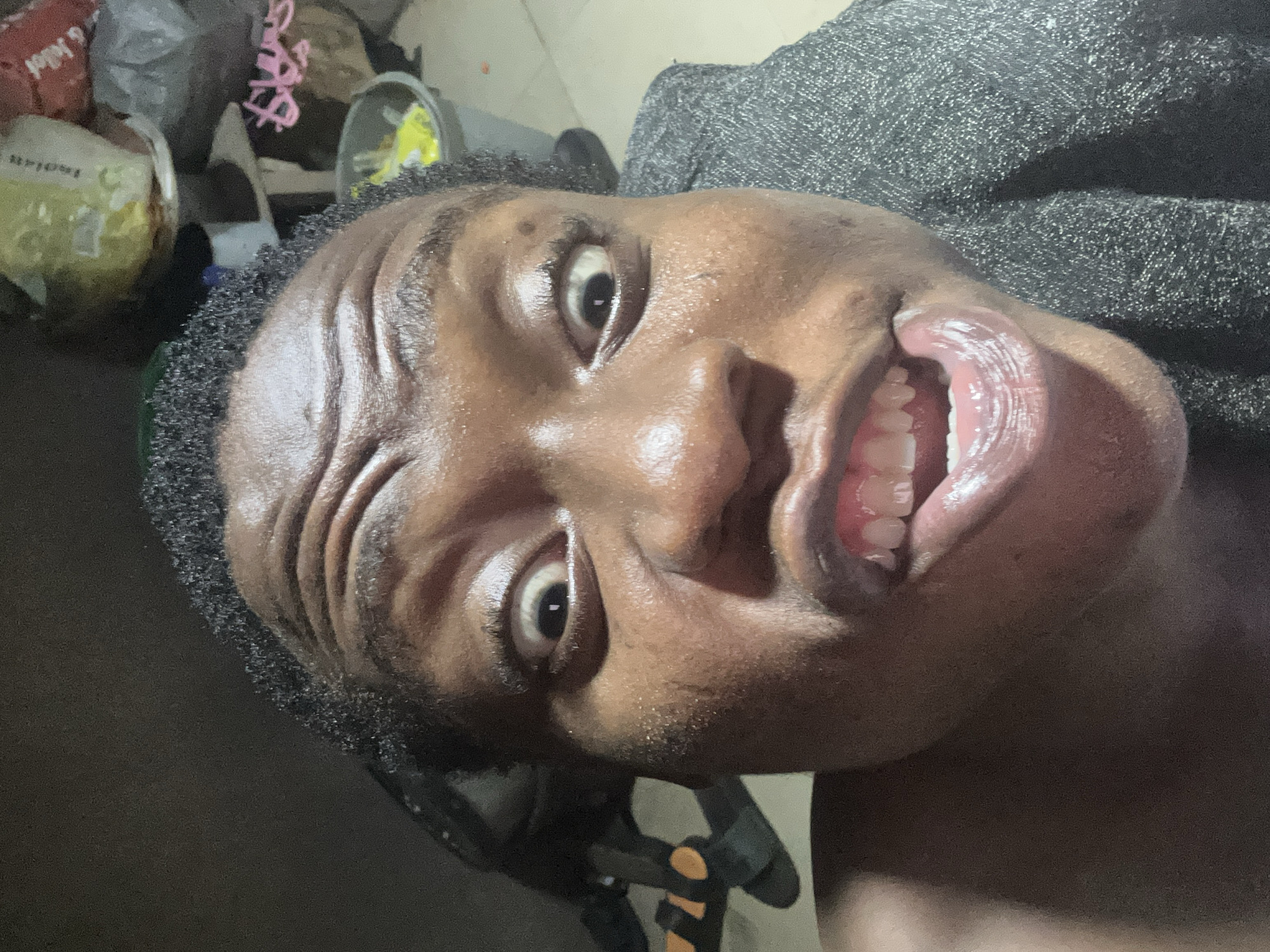 A close-up, slightly angled portrait of a dark-skinned person with short, dark hair, exposing their face and upper torso. Their mouth is wide open, revealing white teeth. The background includes a gray, textured surface, possibly fabric, and miscellaneous items such as bags and containers are seen slightly out of focus. The location and city are not discernible from the image.