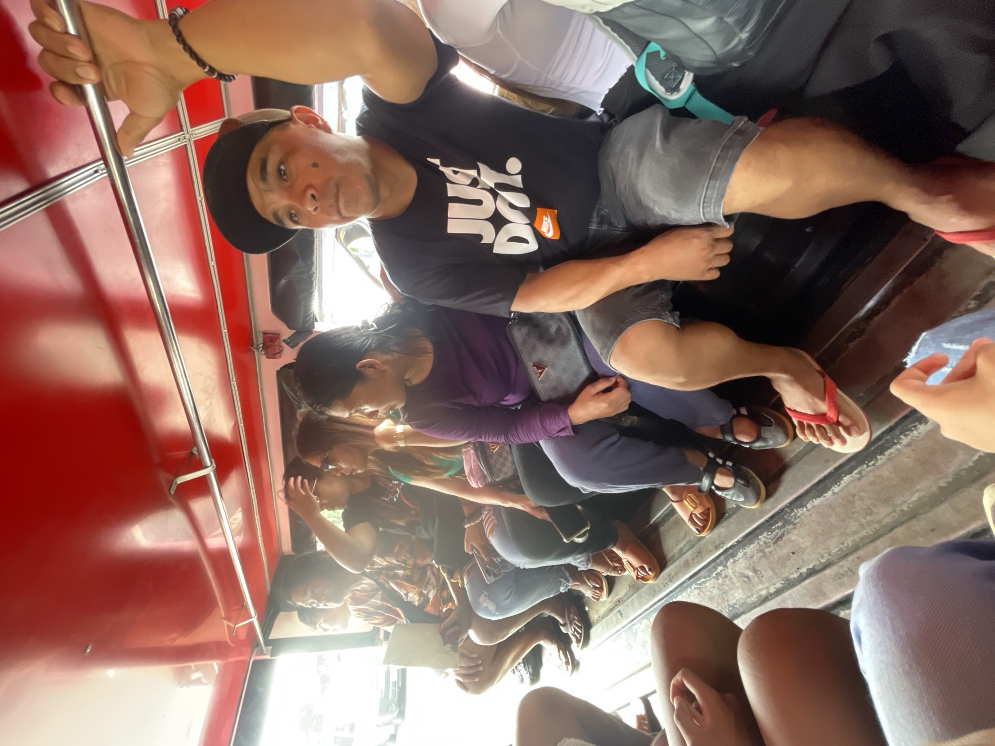The image displays the interior of a public utility jeepney in Davao City, Philippines, characterized by a vibrant red ceiling, two parallel silver grab bars, and a ribbed metal floor. Bright natural light illuminates the scene from the vehicle's left side, revealing a blurred urban environment through the windows.
Seated on the right-side bench in the foreground is a man wearing a black baseball cap, a black t-shirt with "JUST DO IT." and the Nike logo printed in white and orange, and dark gray shorts. He has a distinct dark mark on his left cheek. His right arm is raised, hand gripping one of the silver grab bars, and he wears red flip-flops on his feet. A partial view of another passenger in a white top with a teal strap is visible to his right.
On the opposite bench, a row of passengers is visible. From left to right: A person wearing a patterned short-sleeved shirt, with bare legs and flip-flops, holds a light-colored document and looks towards the left. Next to them, a person with sunglasses and dark clothing has a hand near their head, looking towards the right. Following this, a person with long dark hair, wearing a green top and sunglasses, also looks right; a patterned handbag is on their lap. The last person fully visible on this bench wears a purple long-sleeved top, with a dark patterned bag on their lap, and looks to the right. Their feet are adorned with flat brown sandals.
In the immediate foreground on the left, the lower body of two additional passengers is partially visible; one wears light blue pants, and the other has bare legs. Various footwear, including multiple pairs of flip-flops and sandals, are scattered across the metal floor.
