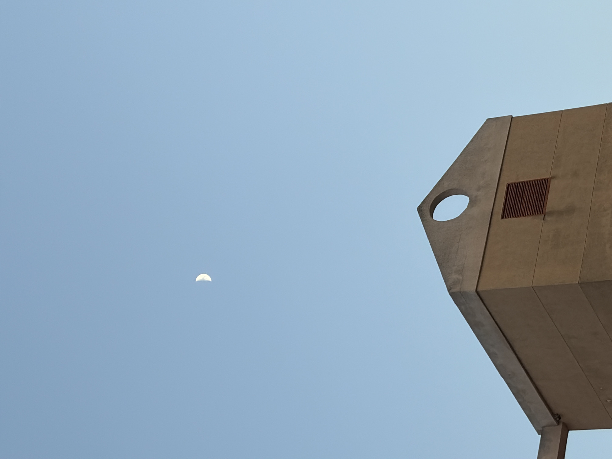 A crescent moon is visible in the clear blue sky above a segment of a beige building in Torrevieja, Spain. The building features a sloped roof with a circular opening at the apex and a dark red rectangular vent below it. The scene is captured from a low angle, emphasizing the vastness of the sky. There are no people or animals present.