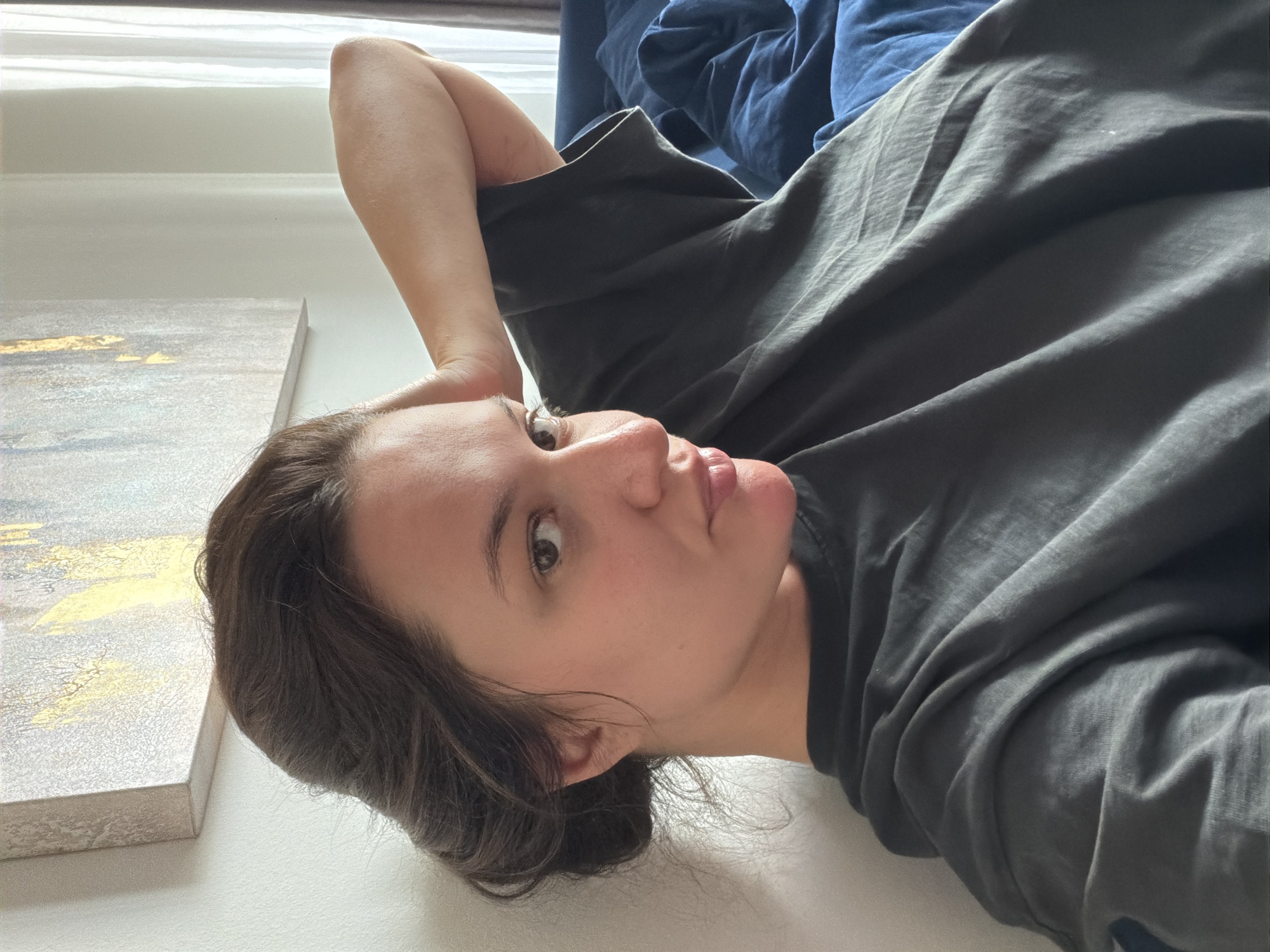 A woman is pictured in a close-up shot, reclining with her left arm behind her head and her right arm bent, resting on her chest. She wears a dark gray t-shirt and has her hair pulled back. The background includes a wall with a framed artwork, a window with light-colored curtains, and a dark blue comforter on a bed. The scene appears to be indoors, possibly in a residential setting in Belgrade, Serbia, given the architectural style and natural light.