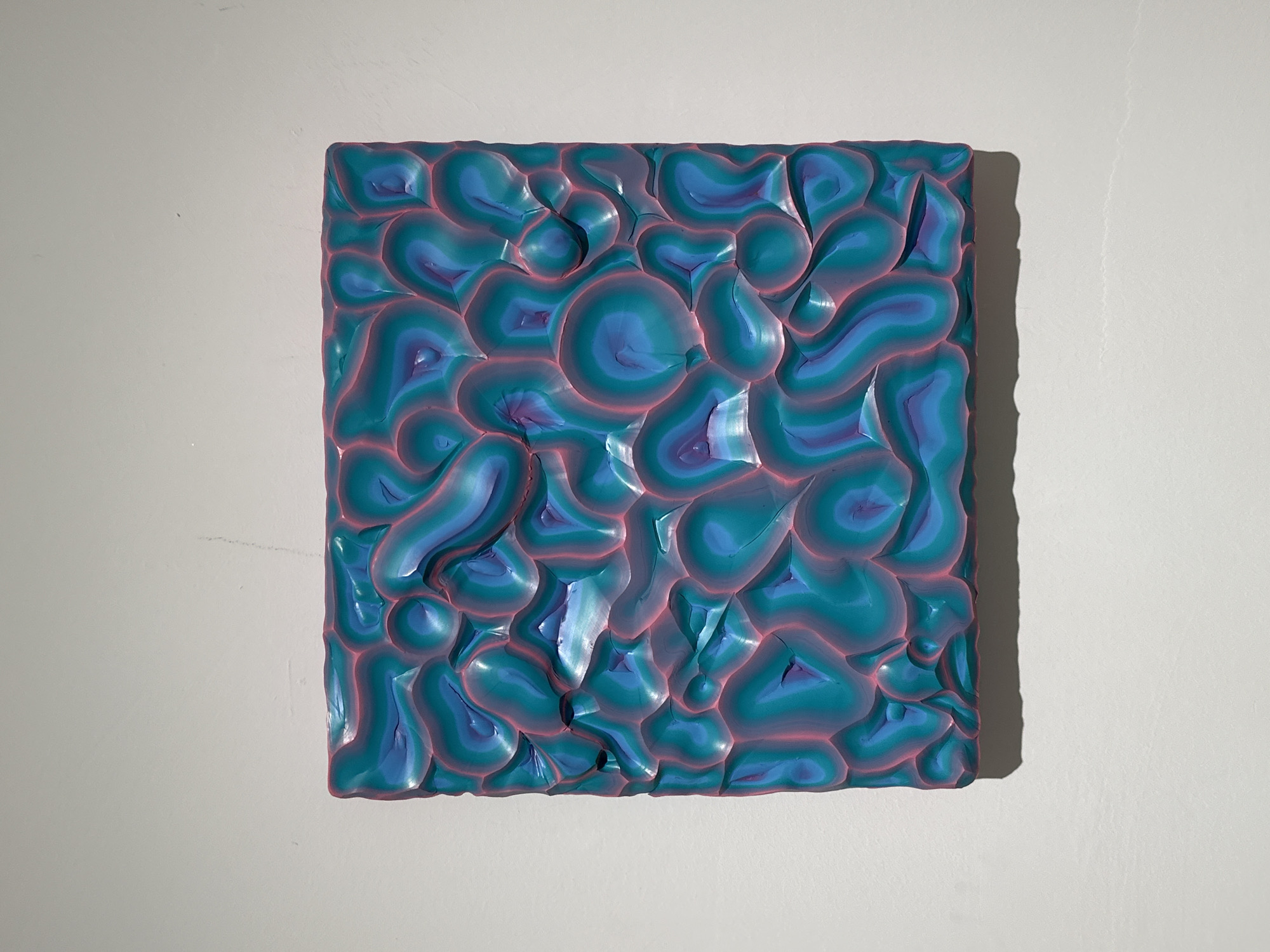 A square, three-dimensional wall art piece hangs on a white wall. The art piece has a textured surface composed of rounded, organic shapes in shades of teal and blue with a thin pink outline. The piece is lit from the side, creating a shadow on the right side of the wall.