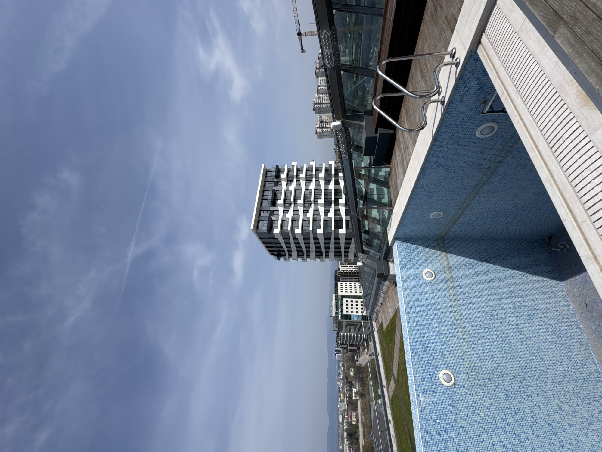 A daytime, elevated view shows a blue-tiled swimming pool, modern buildings, and a clear sky with contrails. The pool's ladder and lights are visible. The buildings feature contemporary architectural designs with balconies and multiple windows. A crane is visible in the distance. The scene depicts a rooftop setting in Sofia, Bulgaria, with a view of the urban landscape.