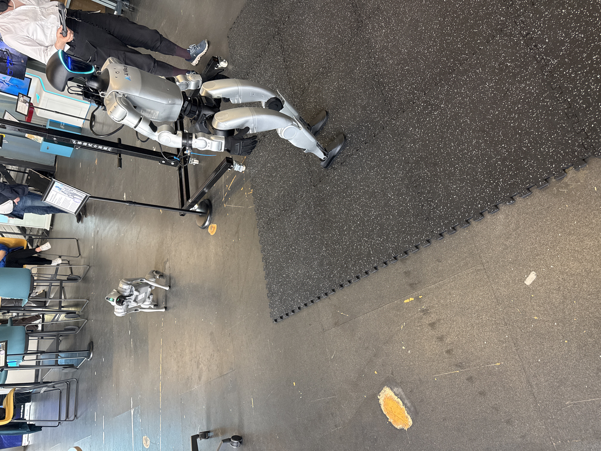 The image displays an indoor scene likely set in a robotics laboratory, exhibition, or demonstration area. The floor consists of dark grey industrial-style flooring, partially covered by black interlocking rubber mats with white speckles. Scuff marks and some light orange-yellow splatters are visible on the grey floor.
The primary subjects are two robots and at least two people. A large, humanoid-like robot, predominantly silver and black, stands on a black support frame. Its head features a black casing with a prominent blue-tinted visor or screen. The text "AI Millage" is visible on its upper torso. Standing near this robot, a person is seen from the waist down, wearing a white shirt, black pants, and grey-blue sneakers. They are holding a black device, possibly a controller or sensor, suggesting interaction with the robot.
Further to the left, on the open floor, a smaller, silver-grey quadrupedal robot is positioned. It resembles a robotic dog, and the alphanumeric "G2" is faintly discernible on its side. In the background on the left, several chairs with metal frames and blue or yellow seats are arranged. The legs of another person are visible, seated. A black display stand with a white information board, which includes a diagram of a robot, is situated near the main humanoid robot.
The environment is brightly and evenly lit, characteristic of indoor lighting. No specific time of day or external weather conditions are evident.