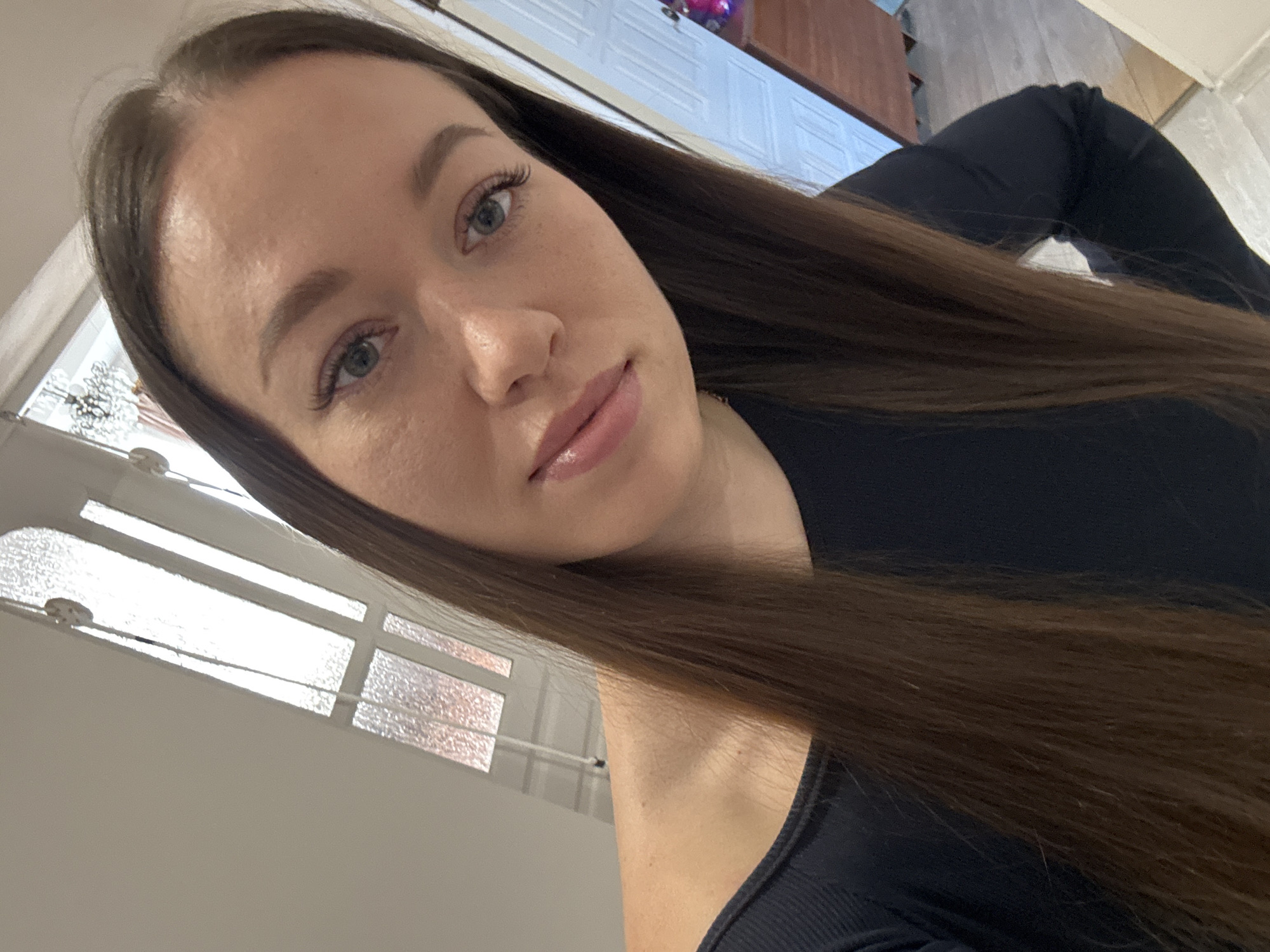 A young woman with long brown hair and light eyes poses for a selfie indoors. She is wearing a black top. The background includes a window with a patterned glass pane and a glimpse of a room with a wooden cabinet. It is not possible to confirm the location as Ферма, Russia, from the image.