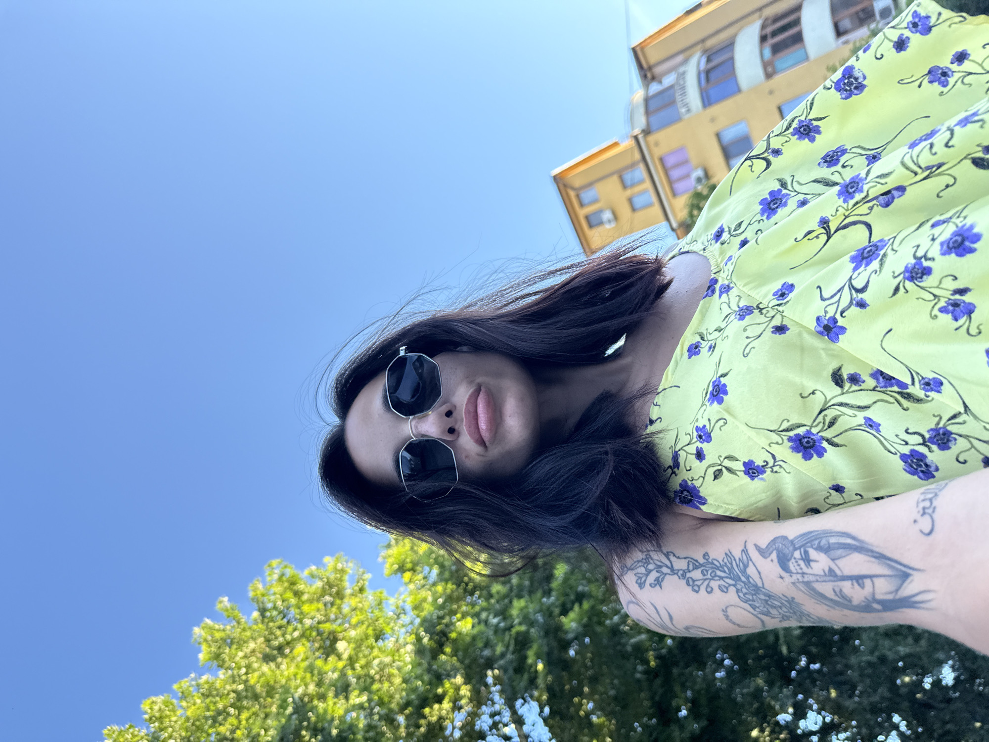 A low-angle, close-up, outdoor, waist-up shot captures a woman with dark hair and sunglasses, wearing a yellow dress with a floral pattern. She has tattoos on her left arm. Behind her, a leafy green tree is visible on the left, and a yellow building with windows and balconies stands on the right. The sky above is a clear blue. The scene is set in the city of Pleven, Bulgaria.