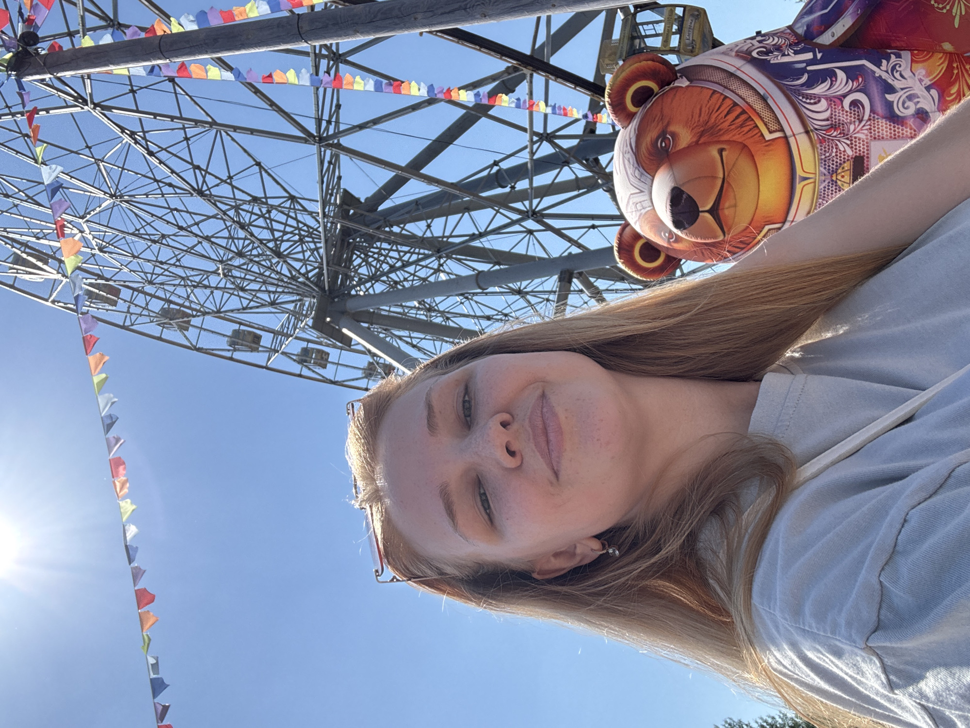 A young Caucasian woman with long blonde hair, wearing a light gray t-shirt and sunglasses pushed up on her head, takes a selfie outdoors. Behind her, a large Ferris wheel is visible against a clear blue sky. A festive string of colorful triangular flags stretches across the sky above the Ferris wheel and a pole draped with similar flags. To the right of the woman, a large, inflatable bear decoration with detailed artwork is positioned. The scene suggests an amusement park or fairground environment, possibly in the city of Shebunino, Russia, indicated by a yellow sign on one of the Ferris wheel cabins that reads "Кострома" (Kostroma).