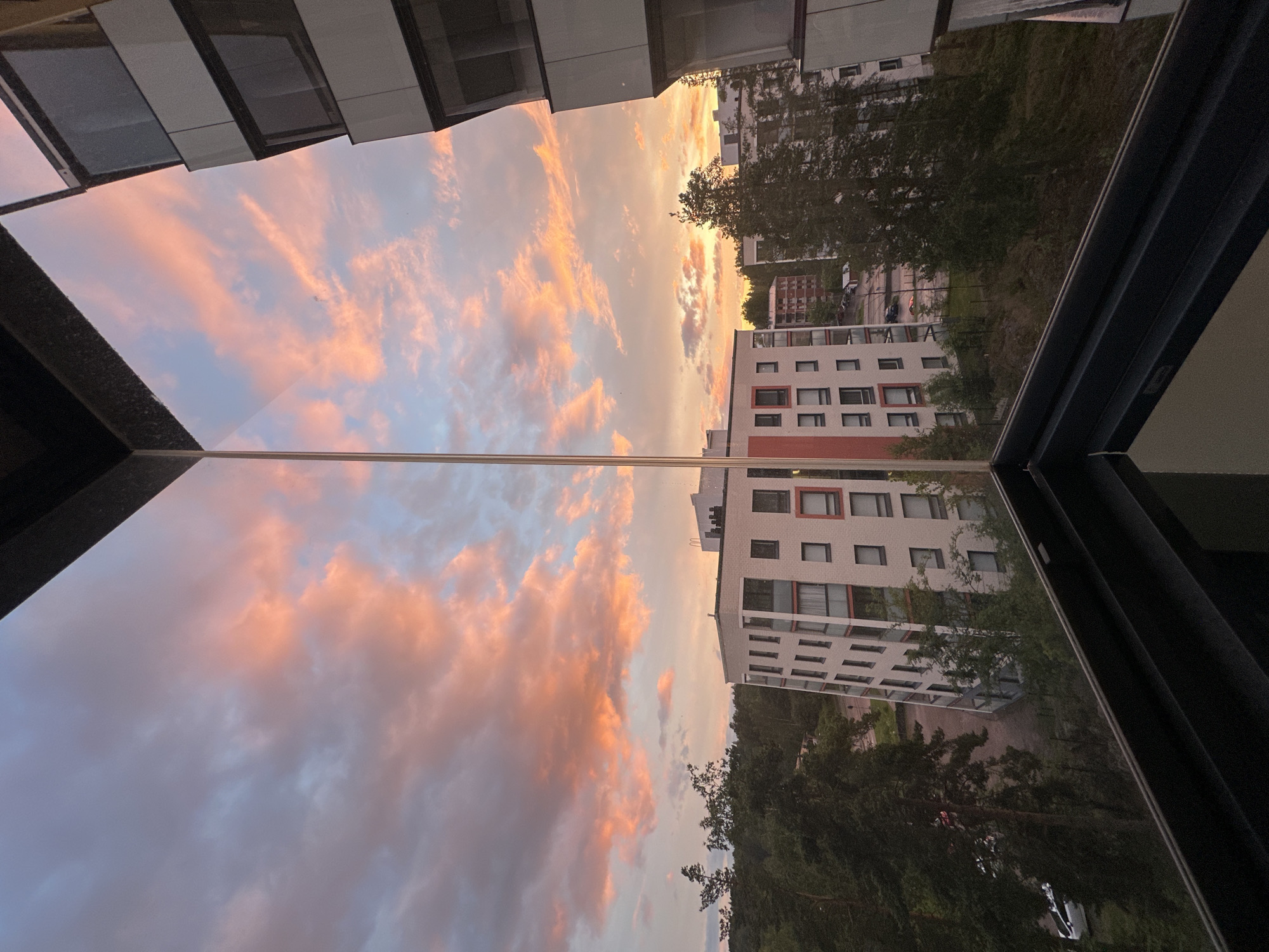 This image captures a reflection of a sunset over a city. The sky is filled with clouds in varying shades of pink, orange, and blue, indicating the time of day is sunset. In the reflection, modern apartment buildings with white and red facades are visible, interspersed with lush green trees. The buildings have numerous windows, some of which are dark, suggesting that some apartments are occupied.
The reflection is captured on a large, glossy surface, likely a window or a balcony railing, which also frames the scene. The angle of the reflection suggests the photograph was taken from an elevated position, looking upwards towards the sky and downwards at the reflected cityscape. There is no visible text in the image. The overall mood is serene and picturesque, highlighting the beauty of the natural sky contrasting with the man-made structures. The scene appears to be in an urban or suburban environment.