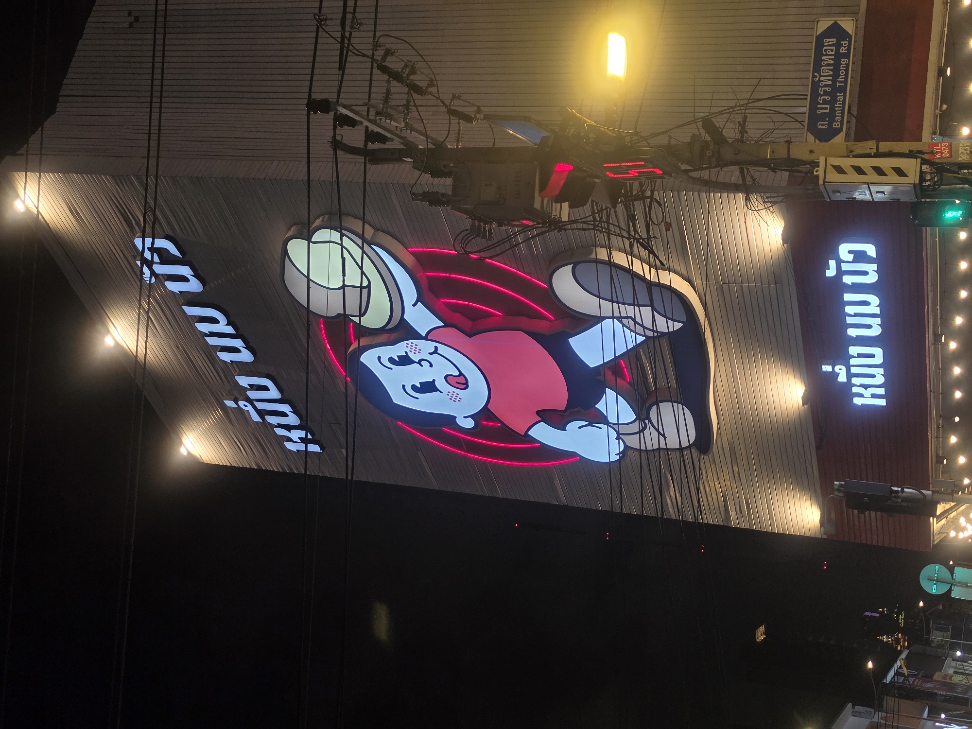 This is a night scene of a building in Bangkok, Thailand. The building has a large, illuminated sign with the Thai words "หนึ่ง นม นัว" (Nung Nom Nua), along with an illustration of a cartoon character. A street sign indicates the location as Banthat Thong Road. There are traffic signals, street lights, and electrical lines visible in the frame. The image is shot from a low angle, emphasizing the height of the building and the signage.