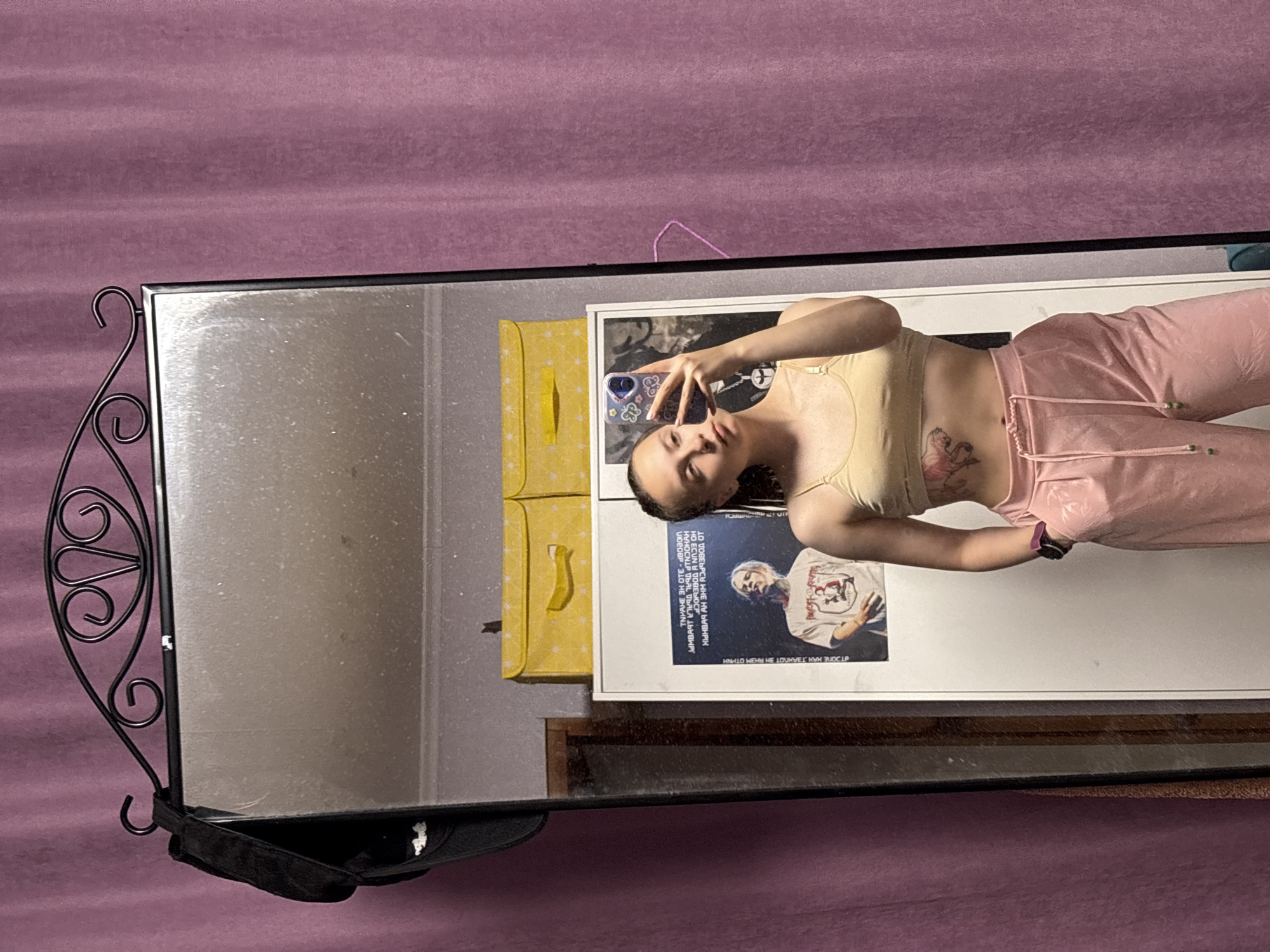 A young woman is taking a mirror selfie with her smartphone. She is wearing a light yellow tank top and pink sweatpants. She has a tattoo on her ribcage. The mirror is a rectangular shape with an ornate wrought iron frame and a small shelf at the bottom. Behind the mirror is a mauve-colored curtain or sheet. A yellow bag or suitcase is visible to the left of the mirror. There's also a poster or print attached to a white surface behind the mirror. The text on the poster seems to be in Russian. The environment appears to be indoors. Прикамский, Russia is not confirmed by visual features within this image.