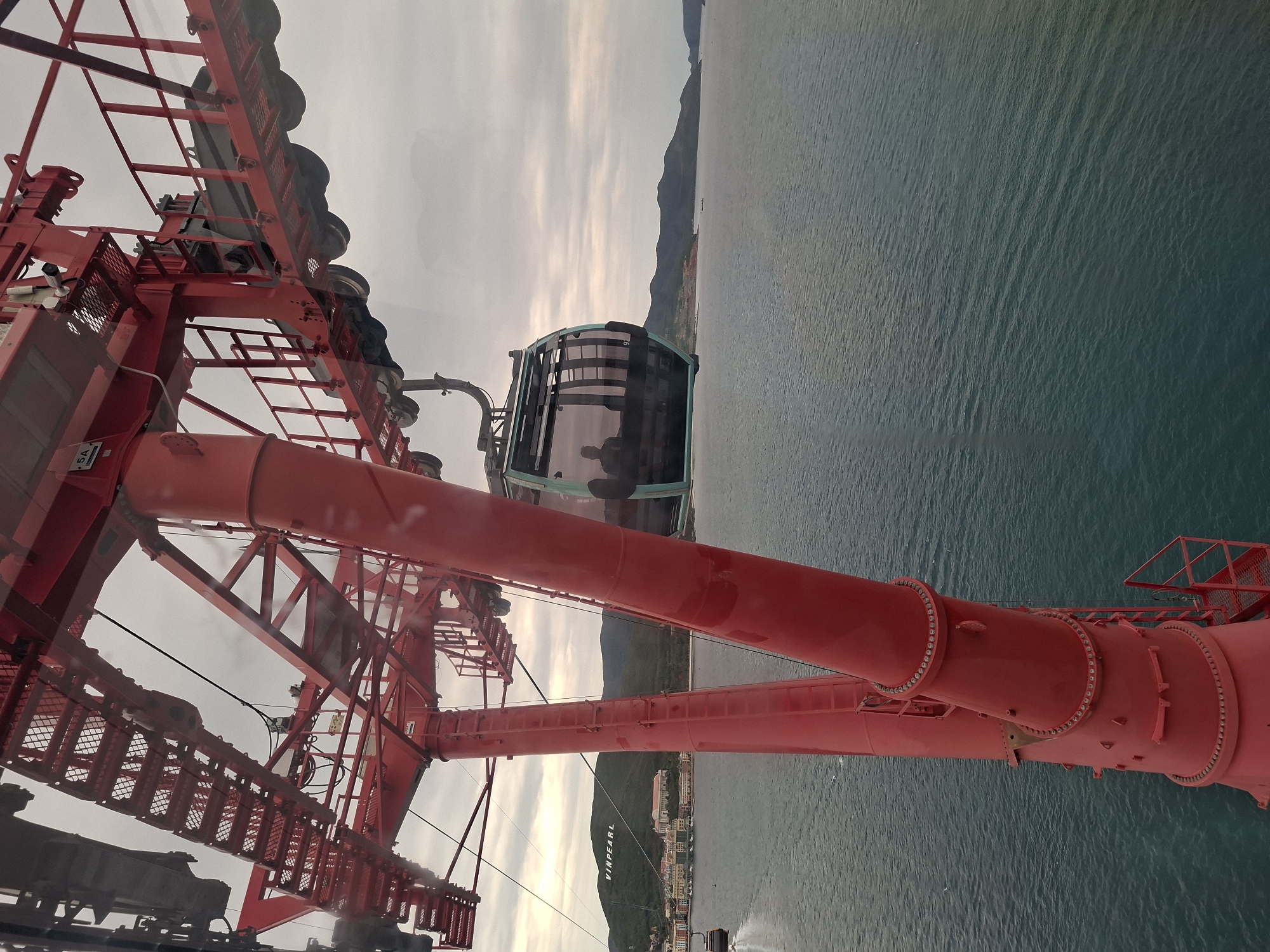 The image, taken from a high vantage point, shows a portion of a large, red cable car tower structure in Nam Nha Trang Ward, Vietnam. A cable car cabin hangs suspended from the cables. The tower's construction is visible, including the framework, supports, and connecting hardware. The sea and coastline are visible in the background, with land and buildings along the shoreline. The sky is overcast.