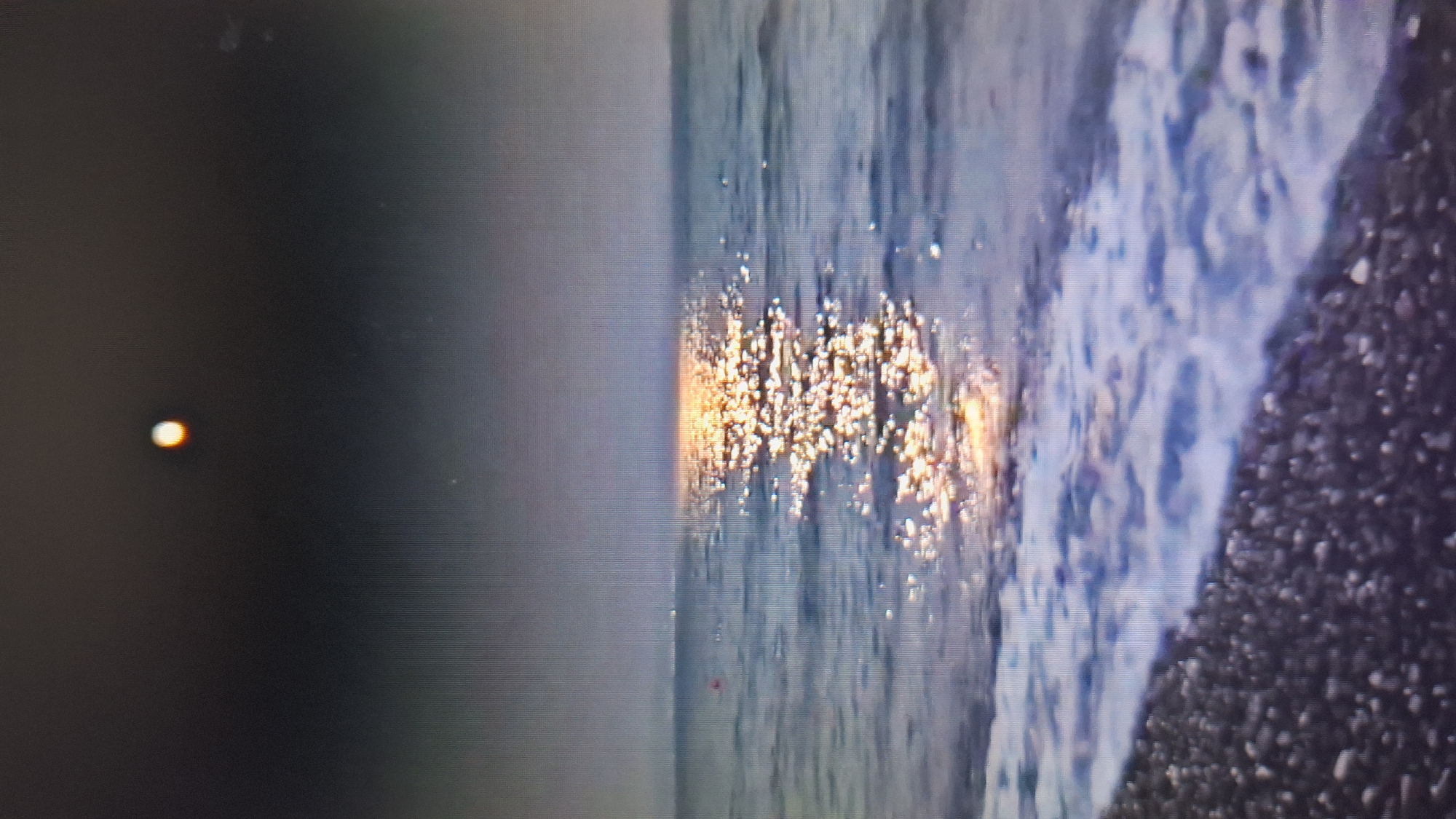 The image, oriented horizontally, captures a tranquil coastal or lakeside scene at dawn, dusk, or under moonlight. In the upper-left corner, a distinct, bright circular orb, possibly the sun or moon, emits a soft orange and white glow against a dark sky.
The central and lower portions of the frame are occupied by a body of water, likely an ocean or large lake. A prominent feature is a dazzling path of sparkling golden and white reflections on the water's surface, indicating a low angle of light from the celestial body. These reflections are numerous and appear like scattered glitter across the water.
To the right, the water meets a shore where gentle waves are breaking. White foam from the breaking waves is visible along the edge of the water. Beyond the water, the shoreline consists of a dark, pebbly or rocky beach, with individual stones discernible.
No people or identifiable man-made objects are visible. There is no visible text. The image's quality suggests it might be a photograph taken of a digital screen, given a subtle pixel grid pattern and some minor dust or imperfections in the darker areas. The scene conveys a sense of peaceful natural beauty, with the interplay of light and water as the primary focus.