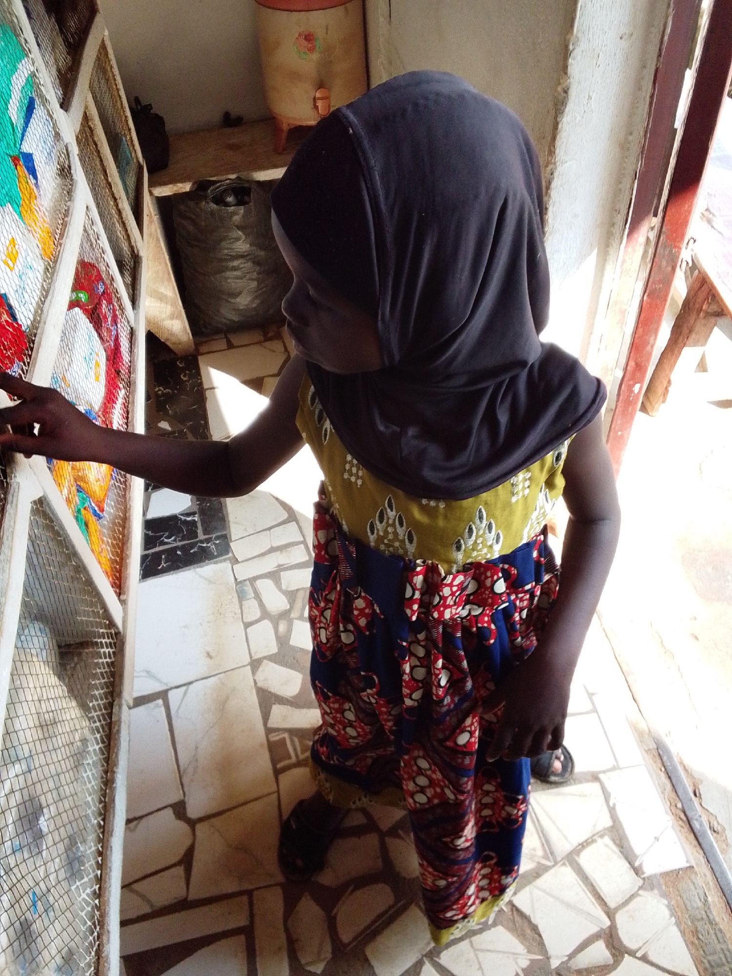 In Serti, Nigeria, a young girl wearing a dark headscarf and a colorful patterned dress is standing near a display of colorful textiles or artwork. The girl is looking towards the display, with her right hand extended and touching it. The display is housed in a wooden frame with a mesh covering. The environment appears to be indoors, with a tiled floor and a white wall. In the background, there are large sacks and a water dispenser. The lighting suggests it is daytime, with sunlight streaming in from the side.