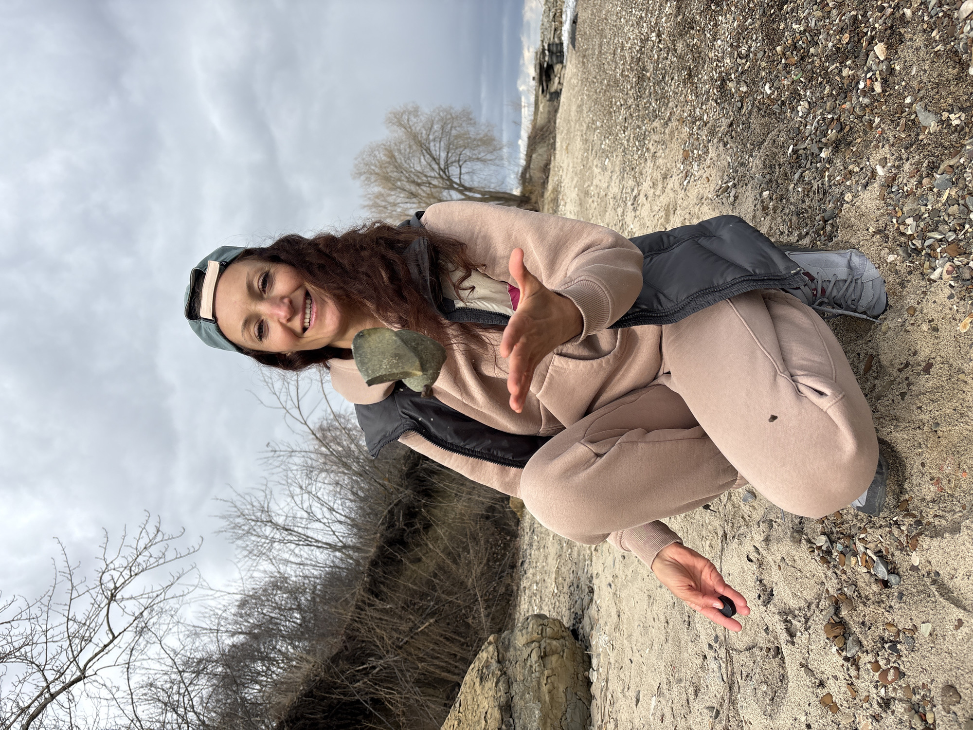 A woman is crouched on a beach, holding a large rock in her left hand and a small, dark stone in her right hand. She is smiling and looking at the camera. She is wearing a pink hoodie and sweatpants, a black vest, a green baseball cap, and gray sneakers. The beach appears to be located on the Volga River, as suggested by the city of Ulyanovsk, Russia. In the background are trees without leaves and a cloudy sky. The ground is a mix of sand and small pebbles.