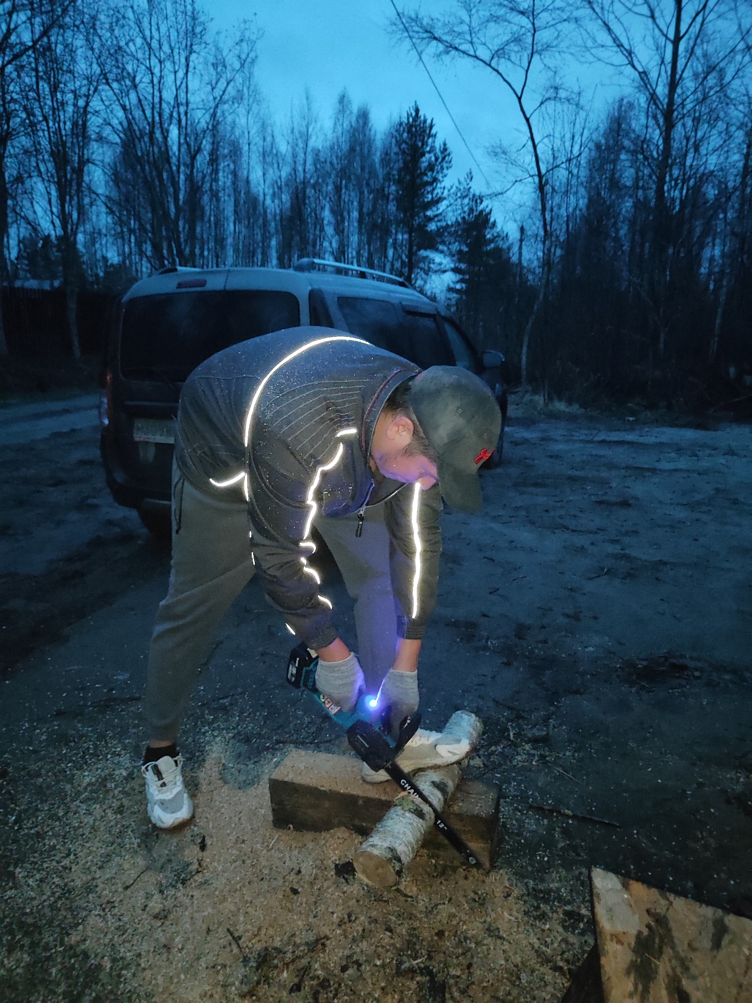 A person is using a chainsaw outdoors at night in Unknown, Unknown. The person is wearing a grey jacket with reflective stripes, grey sweatpants, a baseball cap, and white sneakers. They are cutting a log that is resting on a piece of wood on the ground. There is sawdust on the ground around the log. A vehicle is parked in the background, and trees are visible in the distance. The sky is dark.