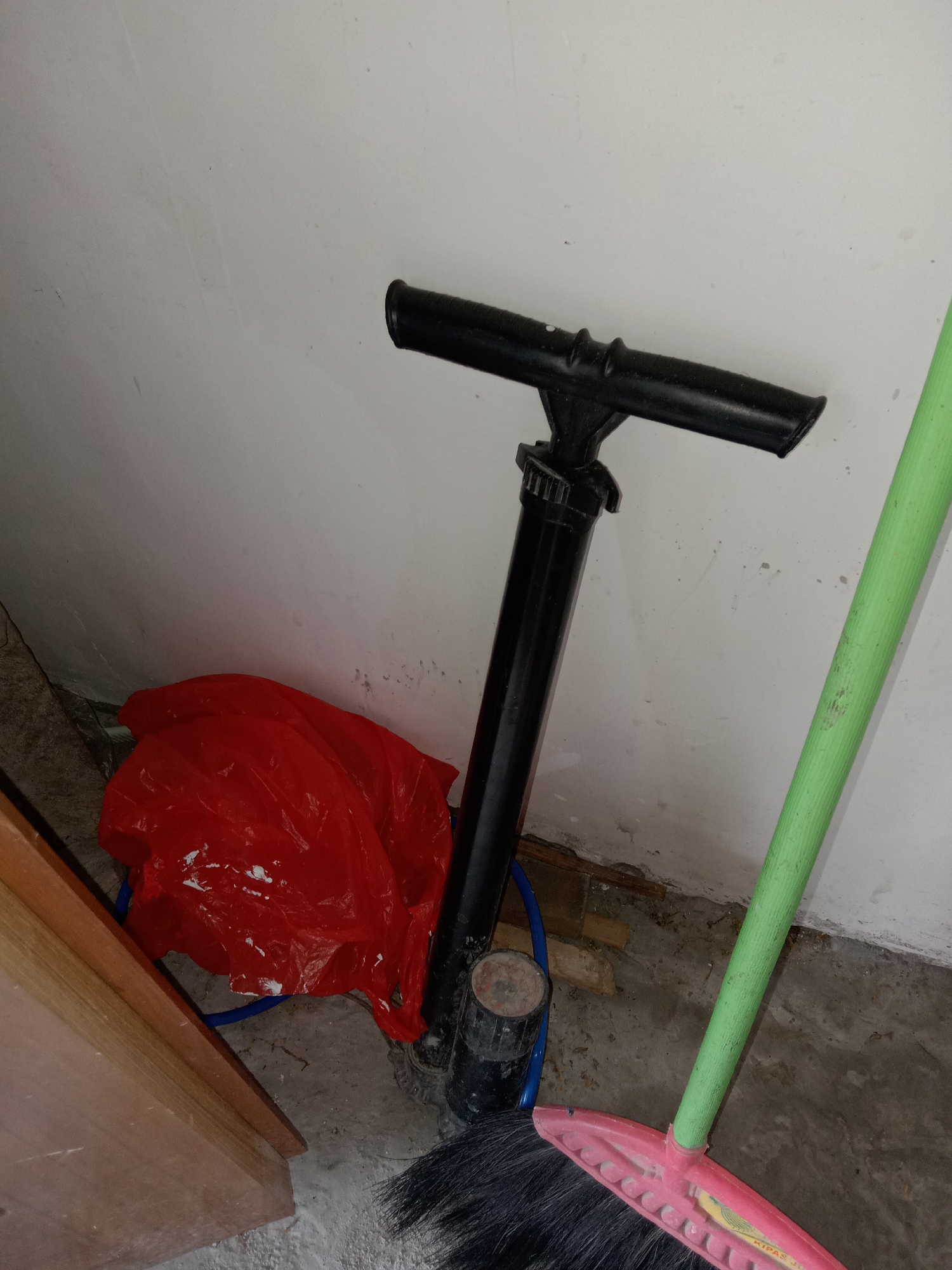 The image depicts an indoor scene, likely a storage area or garage, situated in the city of Pacing, Indonesia. The environment is utilitarian, with a concrete floor and a textured white wall. In the foreground, a black bicycle pump stands upright, its handle extended horizontally. To the right of the pump, a bright green broomstick with a pink brush head is leaning against the wall. To the left of the pump, a crumpled red plastic bag, possibly containing debris or materials, is placed on the floor. A portion of a wooden structure, likely a shelf or cabinet, is visible on the far left edge of the image. The lighting appears to be artificial, casting subtle shadows and highlighting the textures of the objects and surfaces. There are no people present in the image.