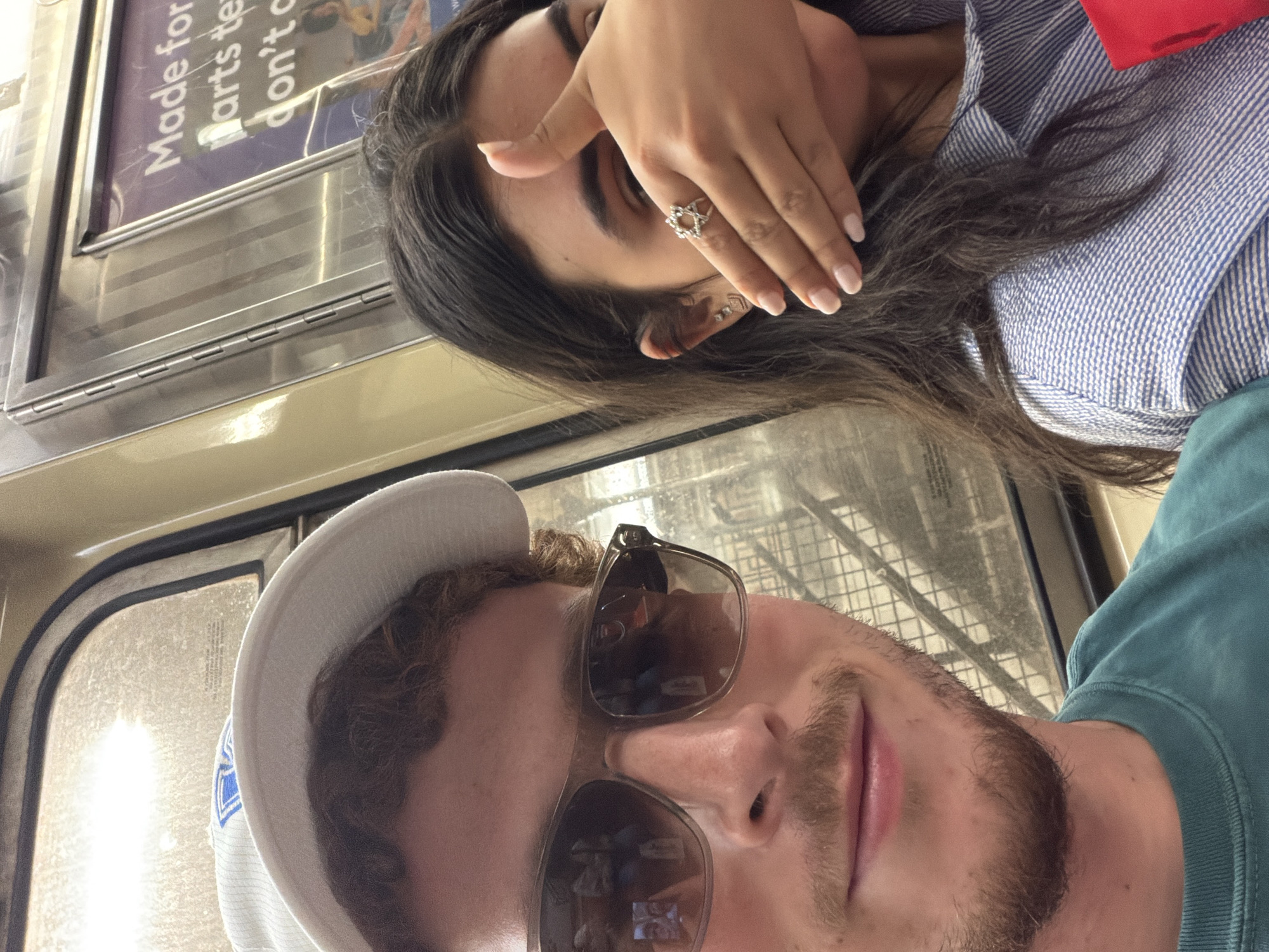 A selfie of two people, a male and a female, possibly in a subway car in New York City, United States. The male has a white baseball cap and sunglasses, a beard, and is wearing a teal t-shirt. The female has dark hair, light skin, and a blue and white striped shirt. She is covering her eyes with her hand. A ring is on her finger. A sign in the background reads "Made for arts te don't". The subway car has a window and metal paneling.