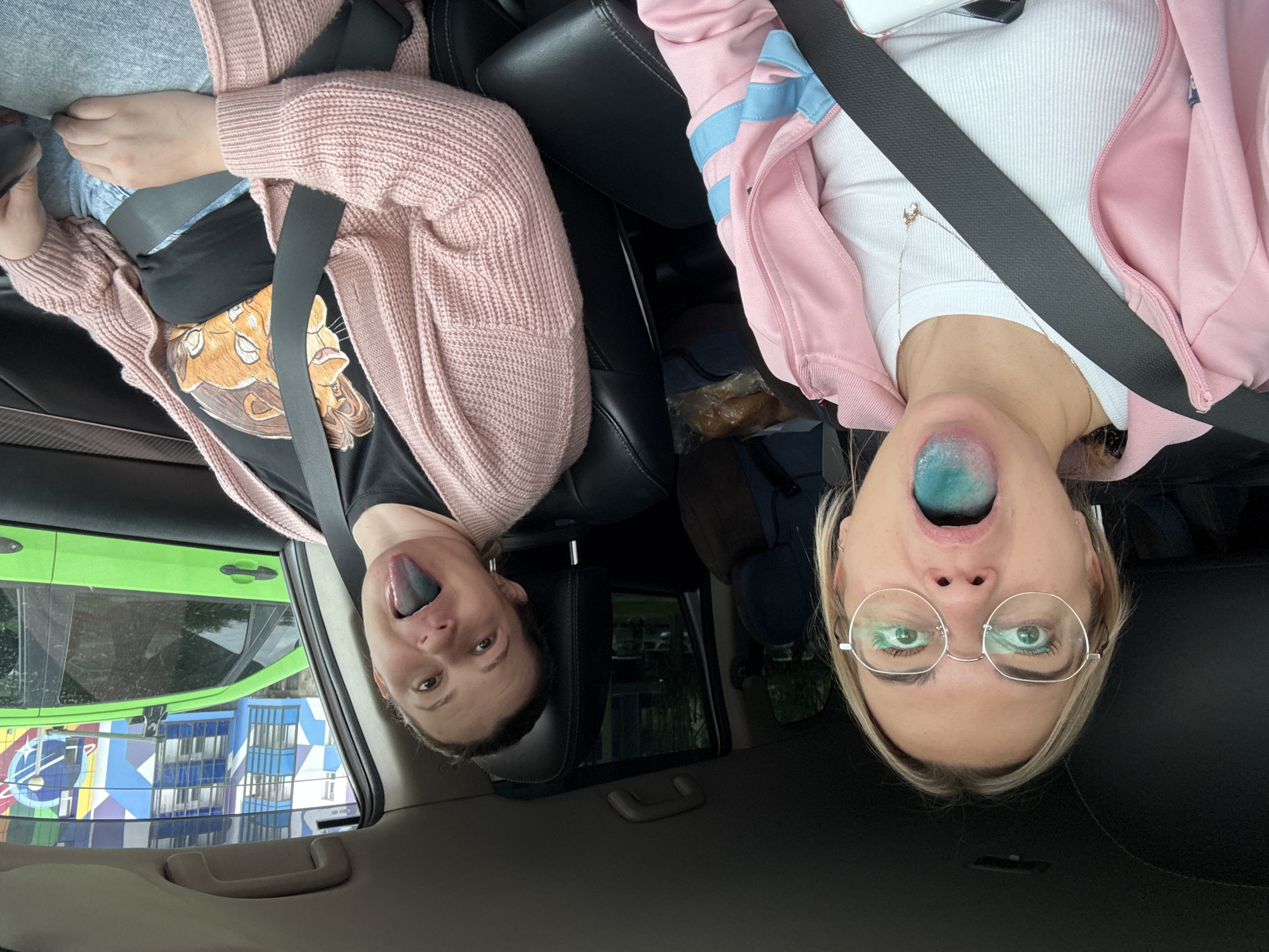 Two women sit inside a car, wearing seatbelts and sticking out their tongues, which are stained blue. The woman on the left wears a black t-shirt with a brown cartoon image, a pink cardigan, and jeans. The woman on the right wears glasses, a white ribbed shirt, and a pink jacket with light blue stripes. Outside the car window is a view of a green car and a colorful building with blue, white, and yellow elements, which is a typical scene in Krasnoyarsk, Russia. The photo appears to be taken inside the car with the camera being upside down.
