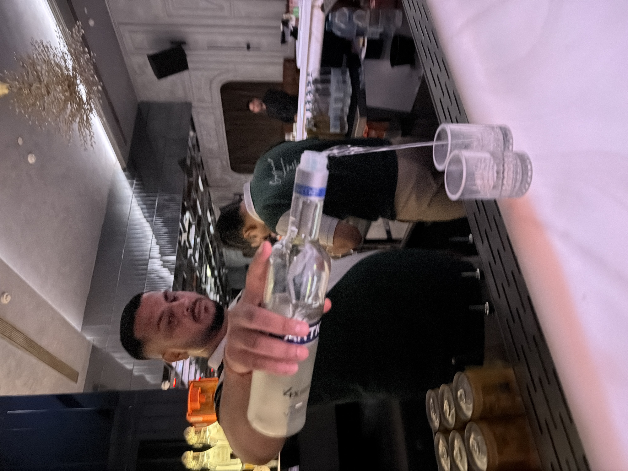 The image depicts an indoor bar scene, located in the city of New Delhi, India. In the immediate foreground, a male bartender with short dark hair and a beard, wearing a dark polo shirt with white trim, is actively pouring a clear liquid from a clear glass bottle into two empty, clear shot glasses positioned on a white bar counter. A stream of liquid is visible flowing into the rightmost glass. The bar counter features a black slotted metal tray directly behind the shot glasses, and several small, gold-colored cans are visible on a shelf to the left of the bartender.
Further into the midground, another male staff member is visible from the back, wearing a dark green polo shirt with white trim and light-colored pants, working behind the bar. Various bottles and bar equipment are arranged on shelves and counters behind him. In the background, partially obscured, are additional patrons; one male figure in a dark shirt is visible standing, and another blurred figure may be seated.
The environment is characterized by modern decor, including a light-colored ceiling with recessed lighting and a decorative, gold-colored, branch-like chandelier hanging on the right side. Walls feature varied textures, including fluted panels on the left and framed paneling on the right. A dark speaker is mounted on the far right wall. The overall lighting is dim with targeted illumination on the bar counter, which glows with a pinkish-purple hue.