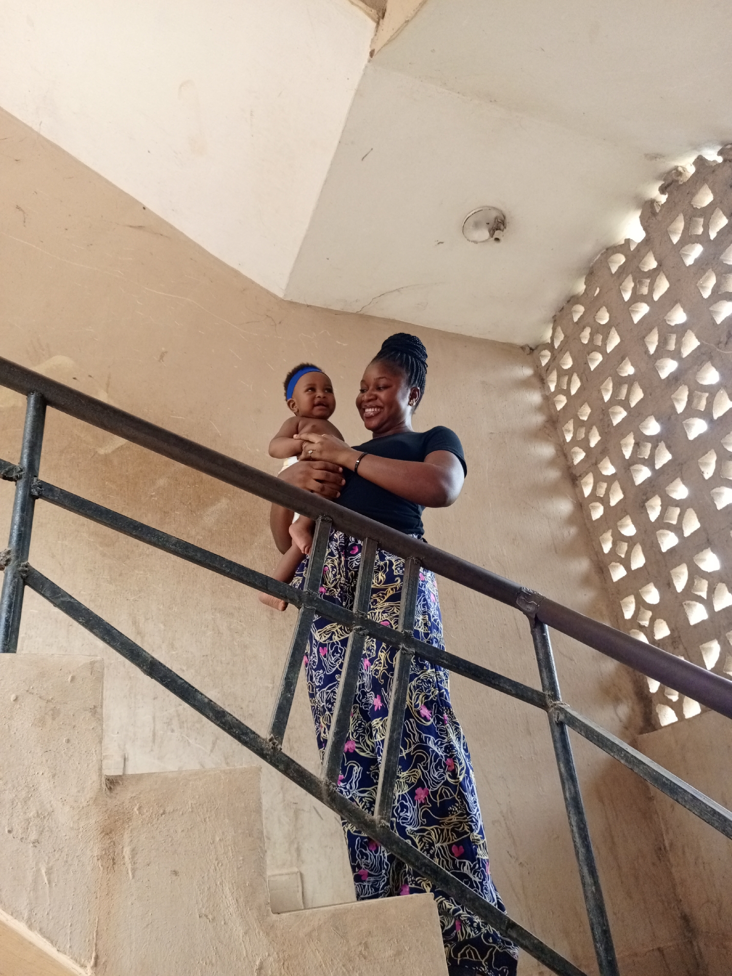 An image captures a joyful moment between a woman and a baby in what appears to be a stairwell or indoor passage. The woman, positioned on a set of concrete stairs, holds the baby in her arms, looking towards the left with a wide, bright smile. She is wearing a black short-sleeved t-shirt and a long, flowing skirt with intricate blue, pink, and gold/yellow patterns. Her hair is styled in braids gathered atop her head.
The baby, held securely by the woman, is wearing a blue headband and a white diaper or cloth wrap, and also appears to be smiling, looking in the same direction as the woman. Their expressions convey happiness and affection.
The setting consists of light-colored, possibly beige or tan, concrete walls and stairs. A dark, possibly black, metal railing with horizontal and vertical bars runs alongside the stairs, separating the subjects from the foreground. In the background, to the right, is a decorative concrete wall with multiple diamond-shaped cutouts, through which natural light is visible, suggesting it's daytime. A simple light fixture is mounted on the ceiling above them. The overall environment appears functional and somewhat rustic, characteristic of an older building. There is no visible text in the image. The scene takes place in Keffi, Nigeria.