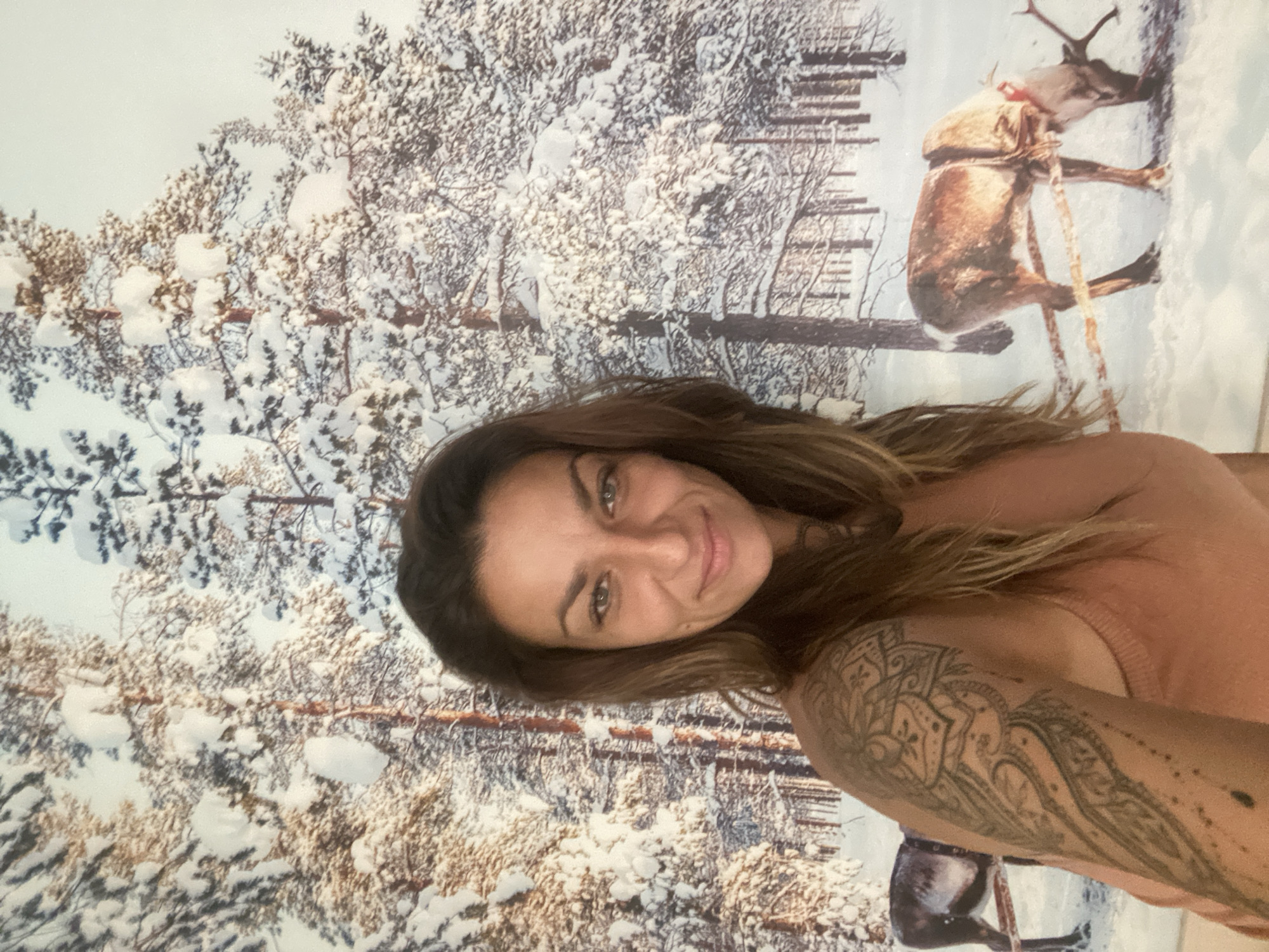 A woman is photographed against a backdrop depicting a winter scene of snow-covered trees and a reindeer with a red scarf. The woman is facing the camera with a slight smile. She has brown hair and is wearing a light pink or peach-colored top. A tattoo is visible on her right arm. The winter backdrop features detailed snowy branches and a single reindeer wearing a harness and red scarf.
There is no indication of the location in the city of Thủ Đức, Vietnam.