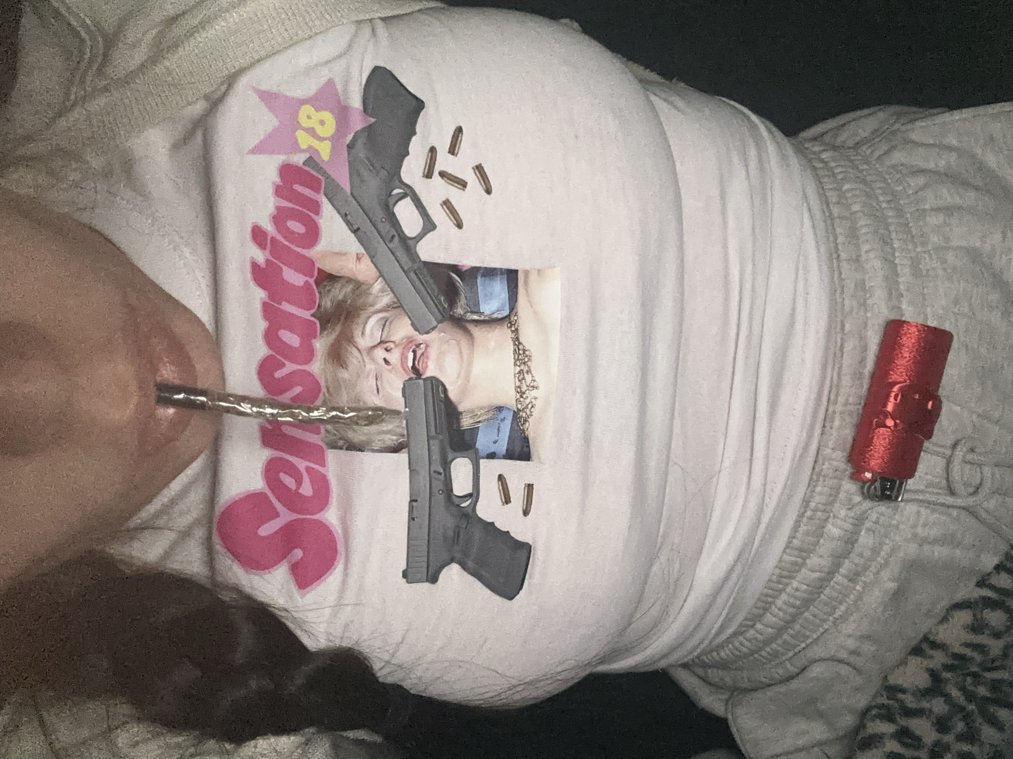 A photo from above depicts a person in bed. The person is wearing a white t-shirt with a print of a woman with two guns pointed at her face and the word "Sensation 18" in pink. There is a metal straw in the person's mouth. To the right of the person is a red lighter and light-colored sweatpants. The location is indeterminable.