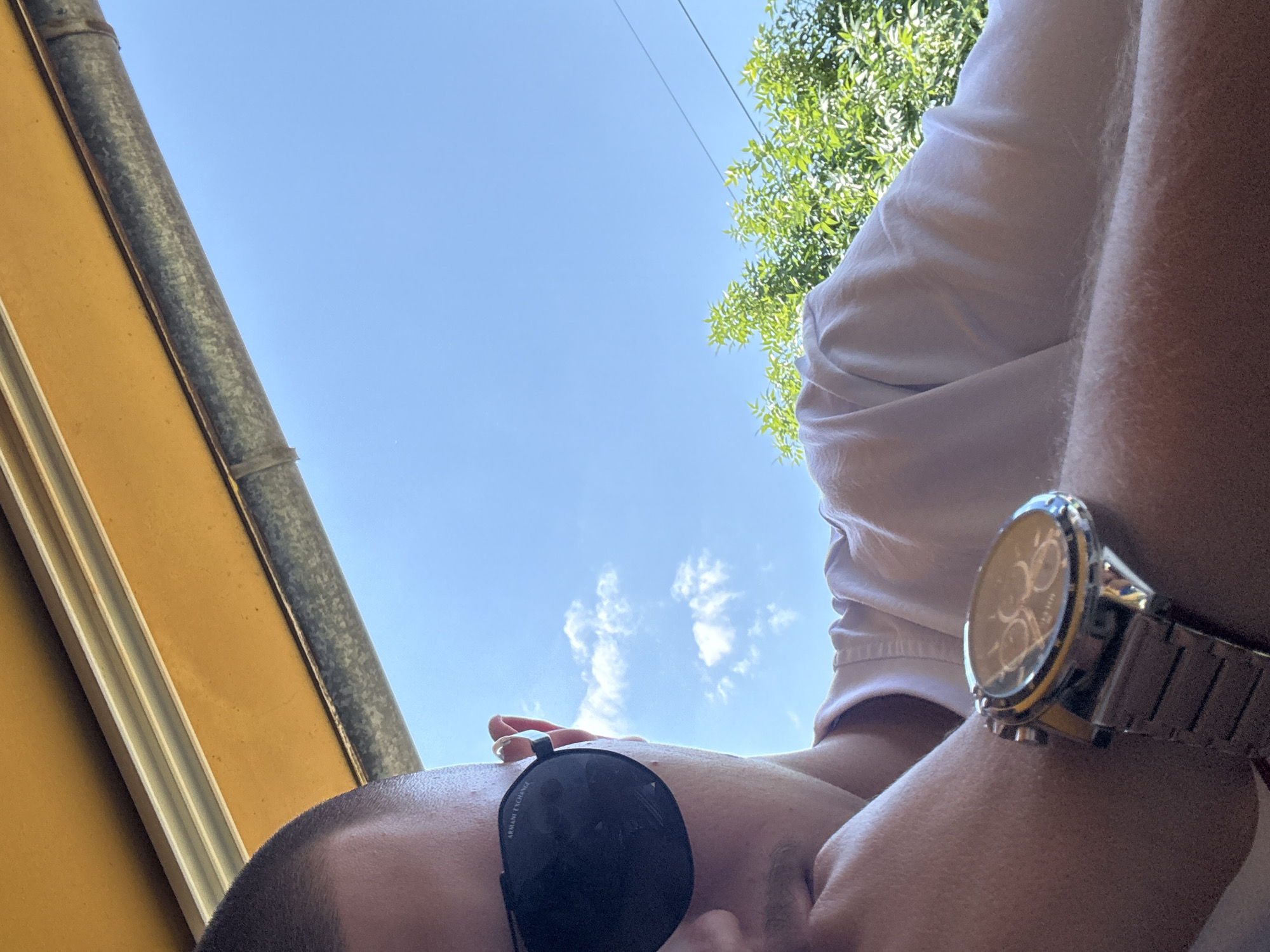 An individual with a closely shaved head and fair skin is captured outdoors, seen from a low-angle perspective, in the city of Blagoevgrad, Bulgaria. The subject is partially obscured, wearing black Armani Exchange sunglasses and a white t-shirt. Their left arm, bearing a metallic watch with a link bracelet and a dark multi-dial face, is visible, with the hand resting near the chin or mouth area. Above the subject, a yellow-orange awning or canopy with visible metallic support beams stretches across the upper frame. The background features a clear blue sky with scattered white cumulus clouds, green foliage from trees or bushes, and thin power lines traversing the upper right quadrant. The scene is brightly illuminated by daylight.