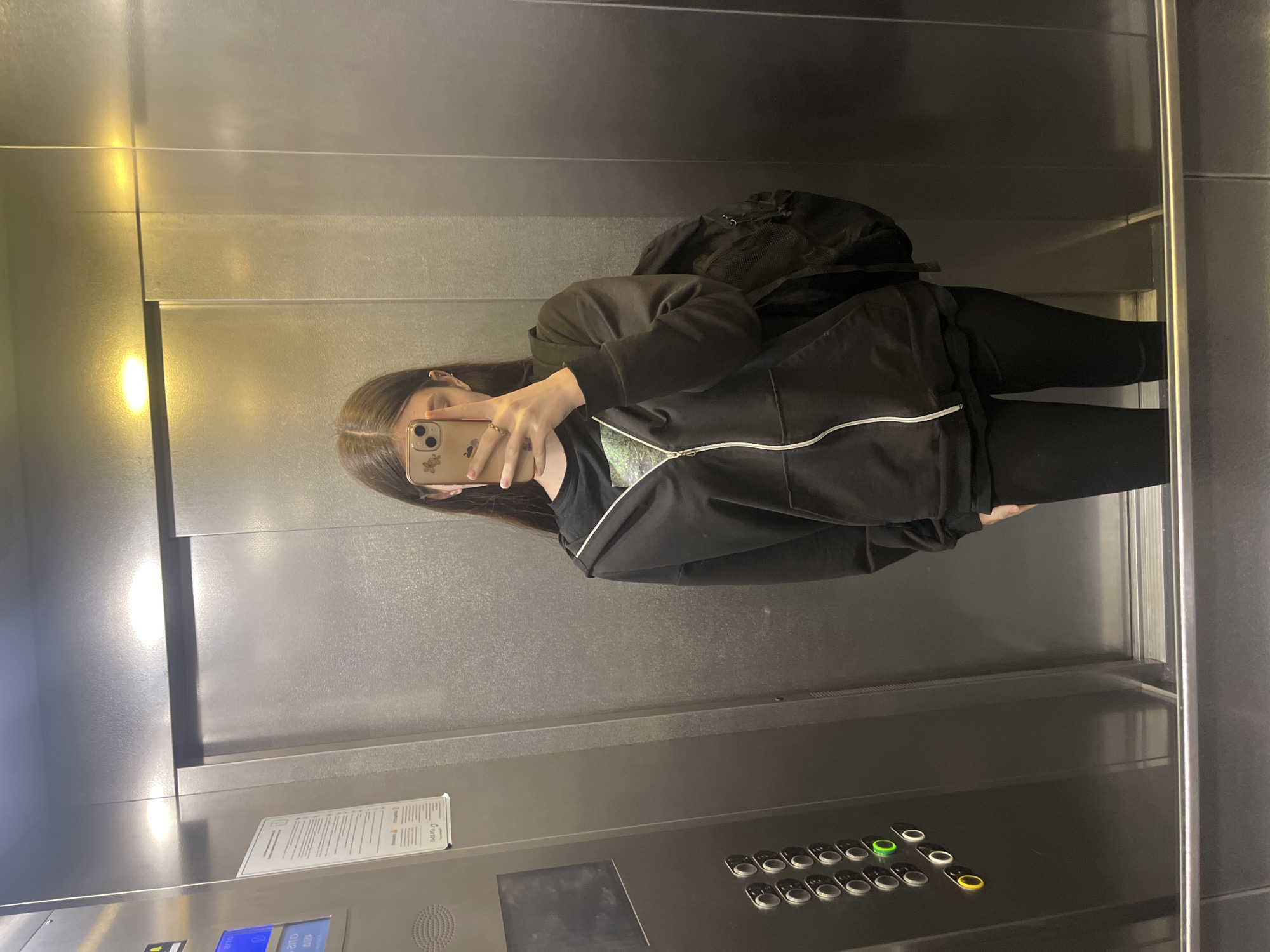 A young person with long brown hair is taking a mirror selfie in an elevator. They are wearing a black zip-up hoodie over a black t-shirt, black leggings, and a black backpack. The elevator walls are metallic and reflective. On the left side of the elevator, there is a panel with buttons, some of which are illuminated. The floor appears to be stainless steel. The lighting in the elevator is overhead and bright, creating reflections on the surfaces. The person's face is partially obscured by their phone as they take the picture. The scene is typical of an indoor, modern elevator.