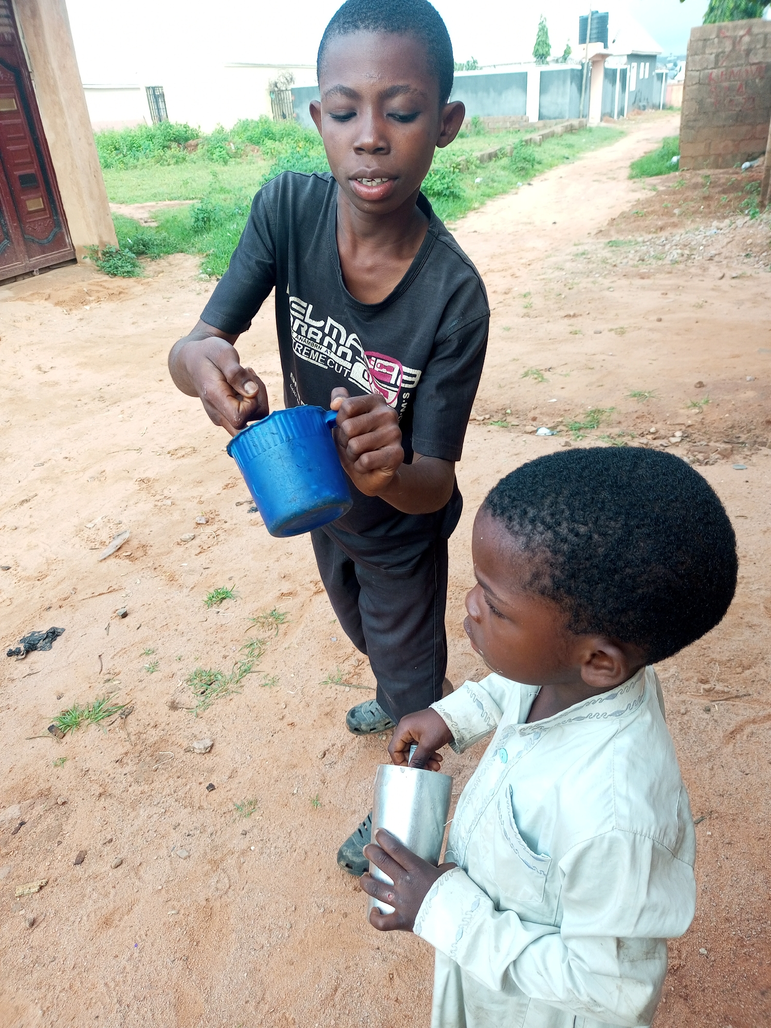 Two young boys stand on a dirt path in an urban area. The taller boy, on the left, wears a black t-shirt and dark pants. He holds a blue plastic cup in his left hand, and with his right hand, he appears to be pouring something into a silver metal cup held by the shorter boy. The shorter boy, on the right, is wearing a light-colored tunic. The background shows a dirt road, some overgrown vegetation, and buildings, including a brick structure and a building with a water tank on its roof. The scene is outdoors, with the ground covered in sand and small stones.