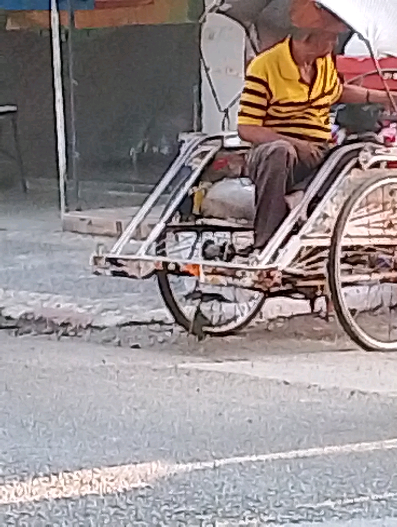 In Probolinggo, Indonesia, a rickshaw driver in a yellow and black striped shirt and brown pants sits in his rickshaw. The rickshaw has a metal frame and a white fabric roof. The driver is partially obscured by the roof and is holding onto the steering mechanism. The rickshaw is on a paved road with a white line marking a lane. In the background, there is a building with a dark, textured wall and a partially visible sign.